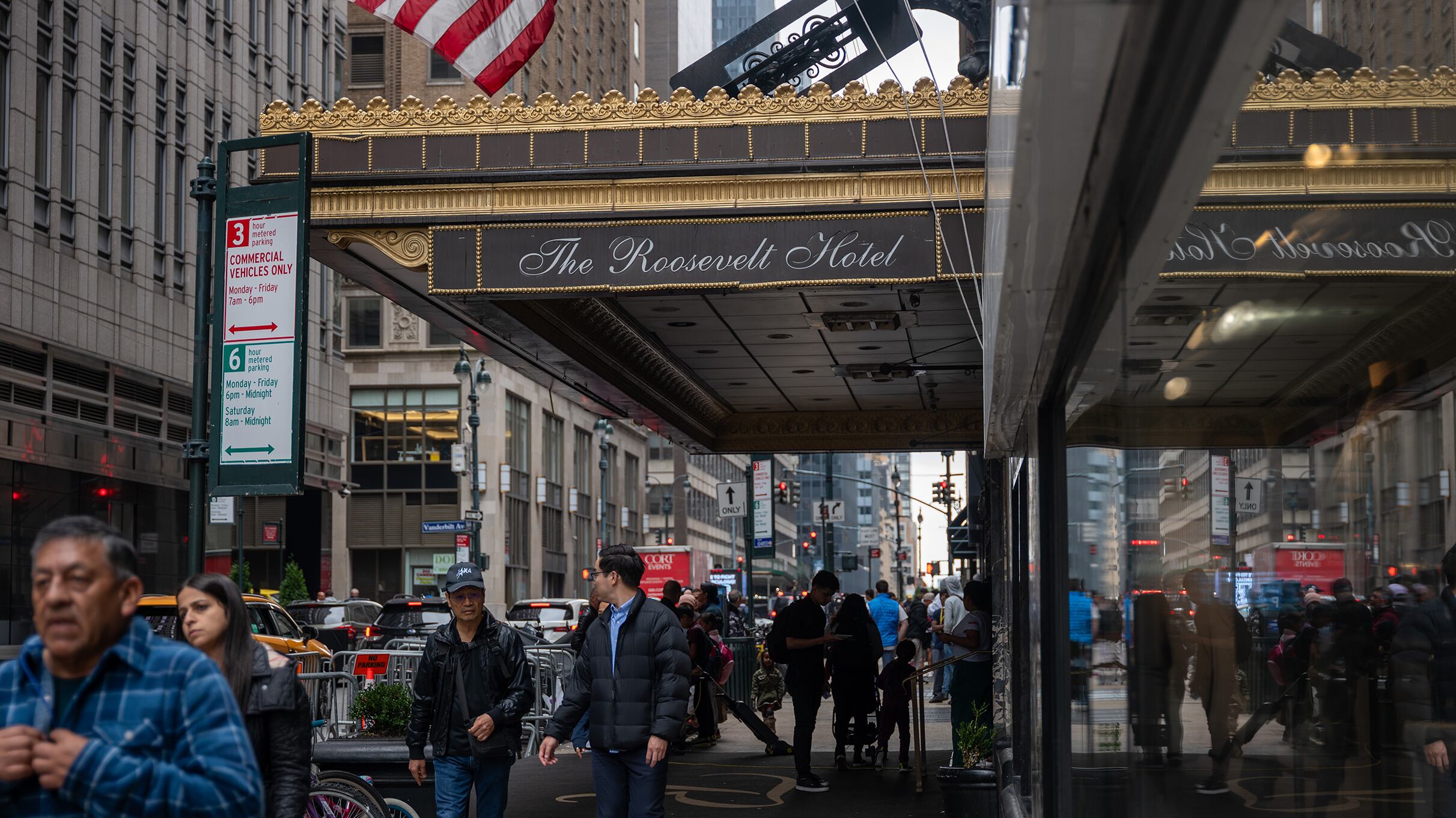 People walk on a sidewalk near a large window of a building with a large overhang with the words " The Roosevelt Hotel."