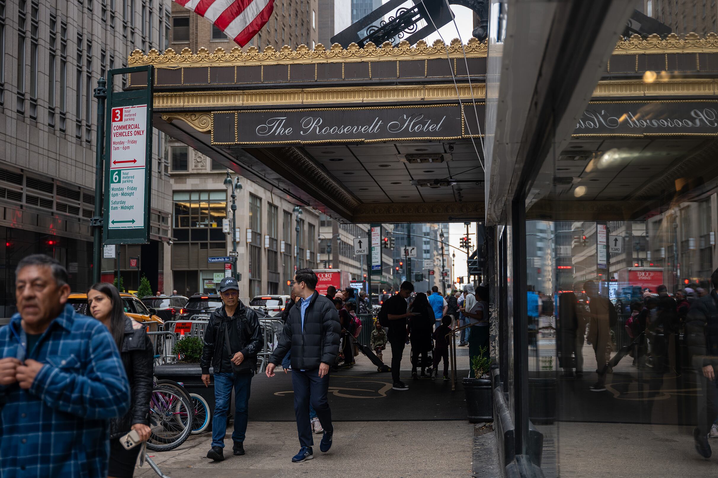 People walk on a sidewalk near a large window of a building with a large overhang with the words " The Roosevelt Hotel."