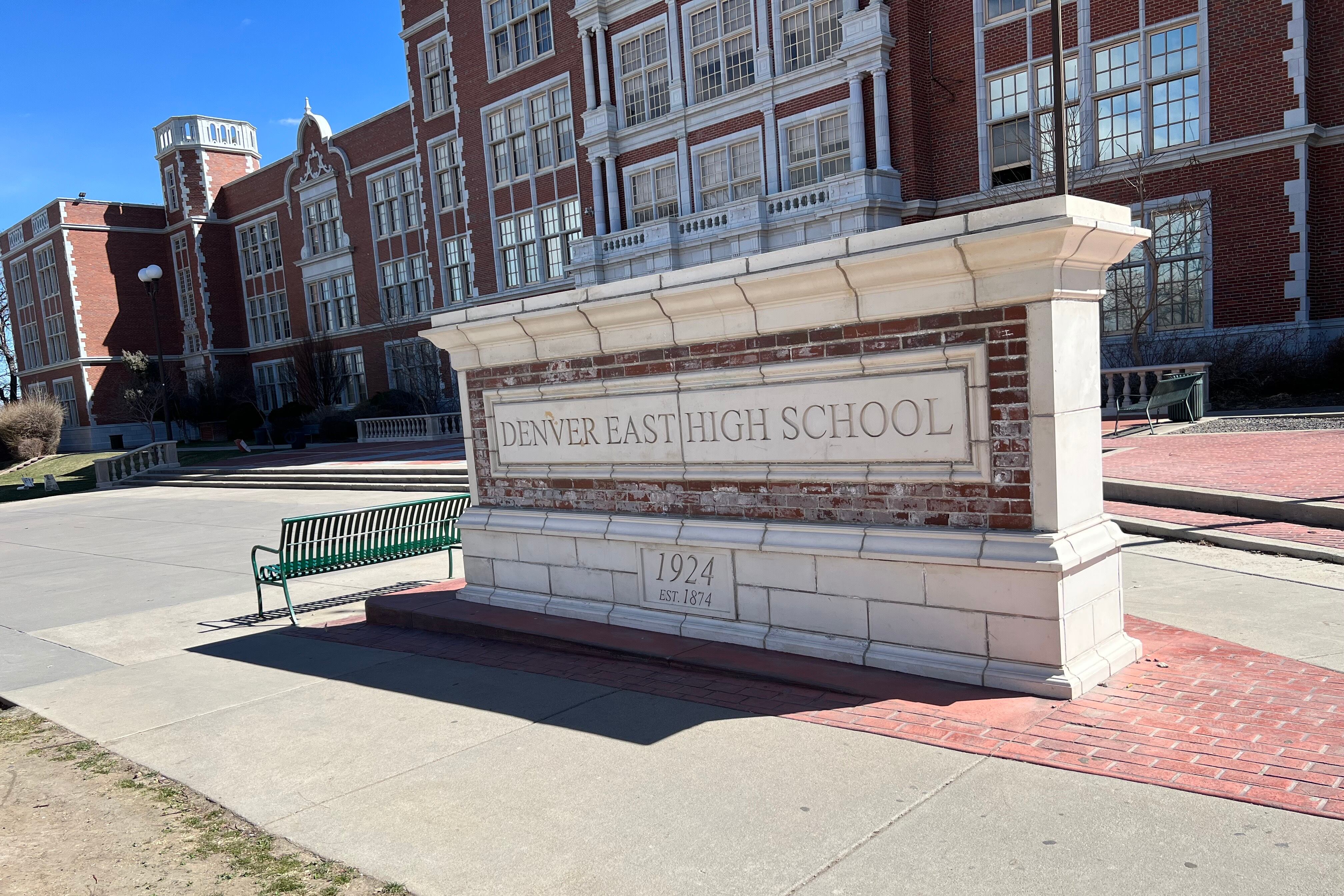 A stone sign that says “Denver East High School” sits outside a brick school building.