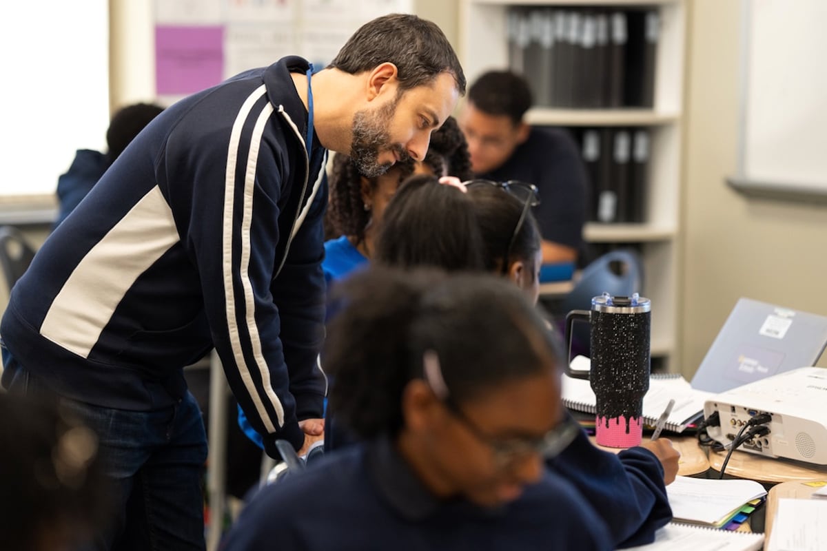 A man in a dark jacket leans over a student at work.
