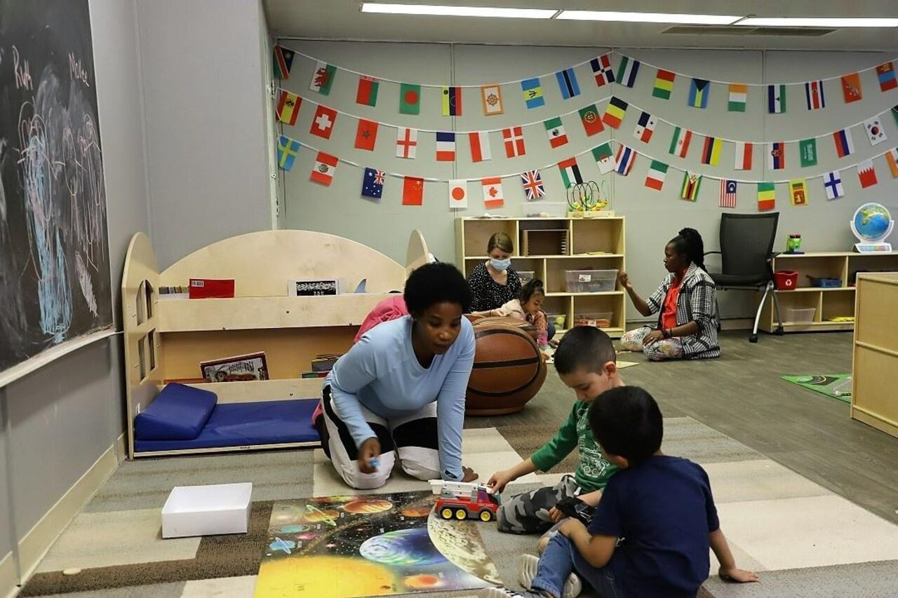 Two women sit on the floor and play with young children in a preschool classroom decorated with flags of many nations. The woman near the front of the image interacts with two boys playing with a truck. The sit around a floor puzzle of the solar system.