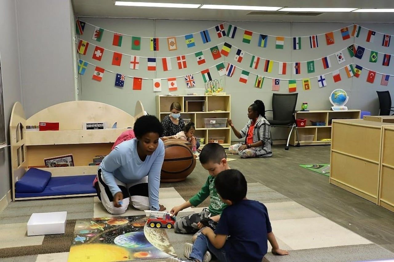 Two women sit on the floor and play with young children in a preschool classroom decorated with flags of many nations. The woman near the front of the image interacts with two boys playing with a truck. The sit around a floor puzzle of the solar system.