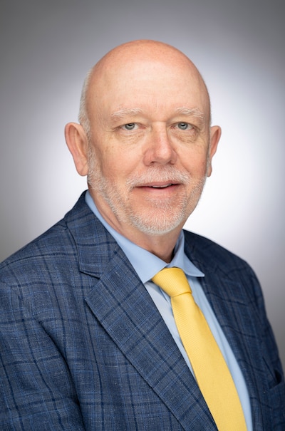 A man wearing a blue suit and a yellow tie poses for a portrait in front of a grey wall.