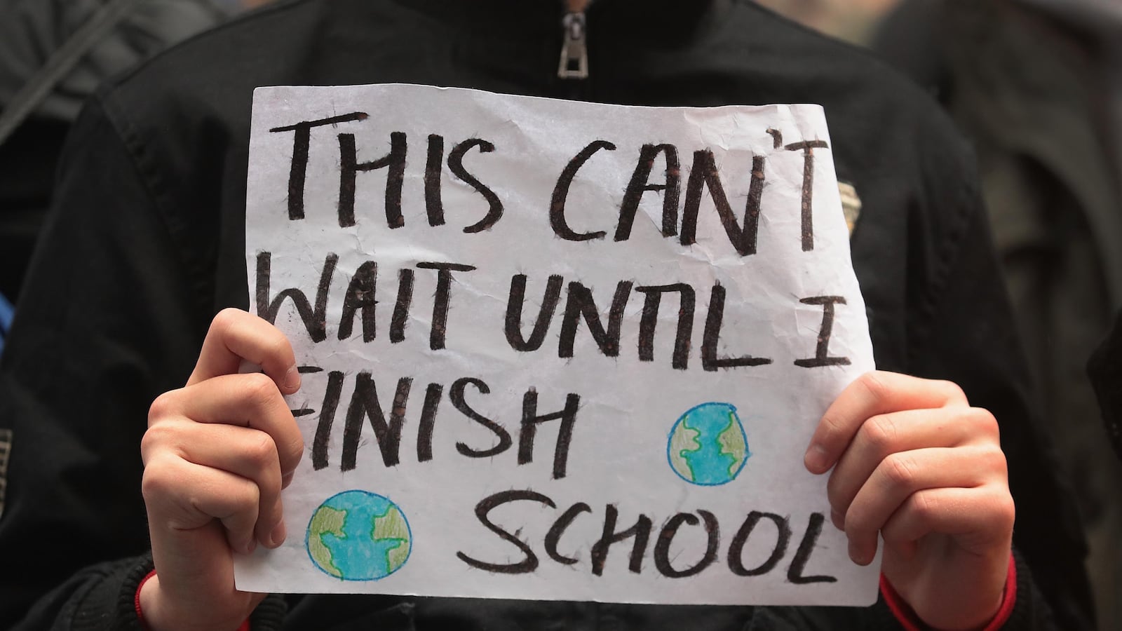 A student holds up a sign that says, “This can’t wait until I finish school.” It has two pictures of the earth on it.
