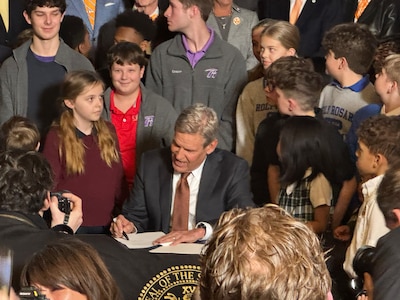 A man in a suit sits at a table signing a piece of paper while surrounded by a group of children.
