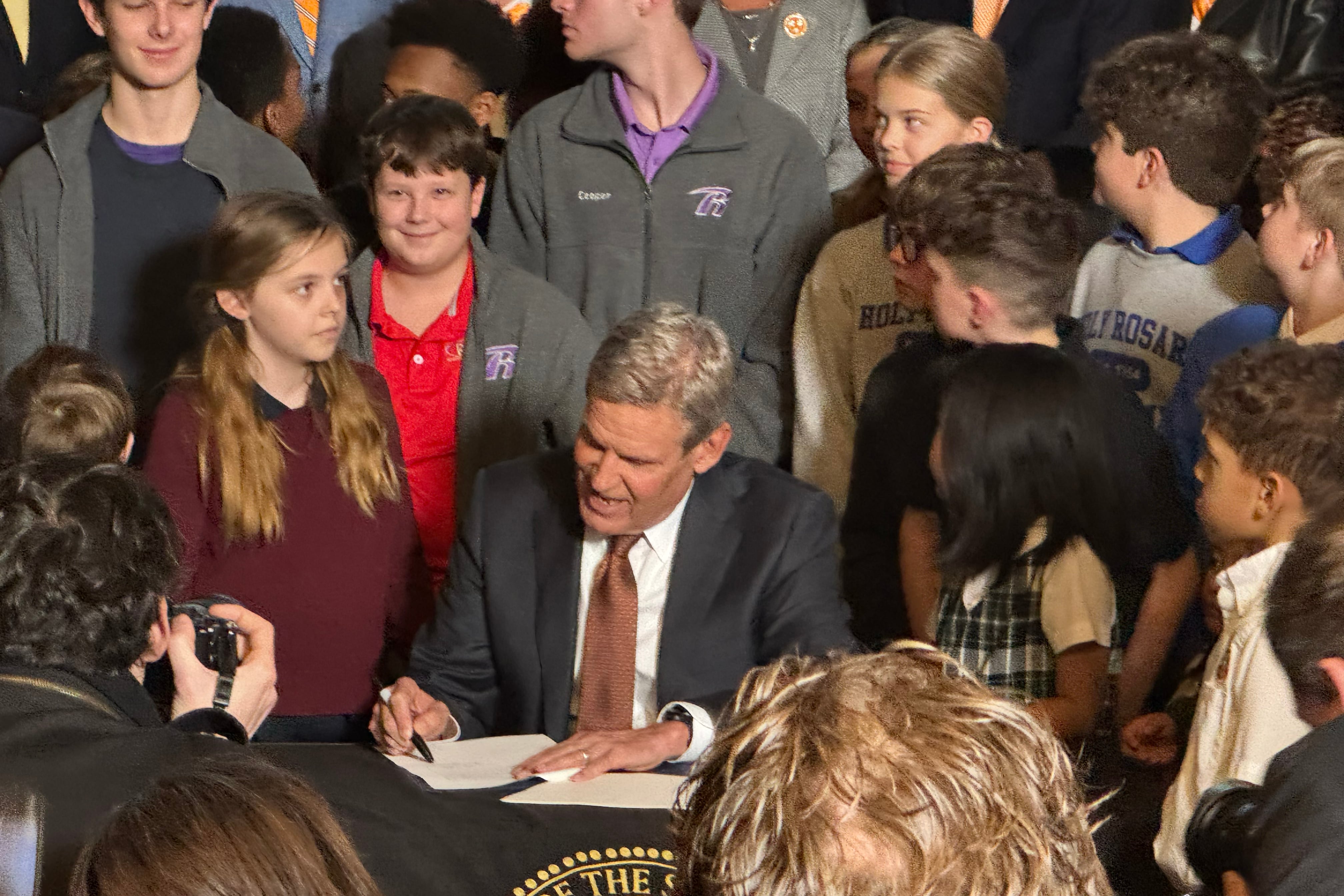 A man in a suit sits at a table signing a piece of paper while surrounded by a group of children.