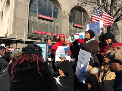 A photograph of adults wearing jackets and holding protest signs outside.