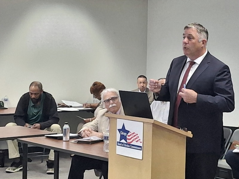 A photograph of a man in a suit standing in front of people in a conference room.