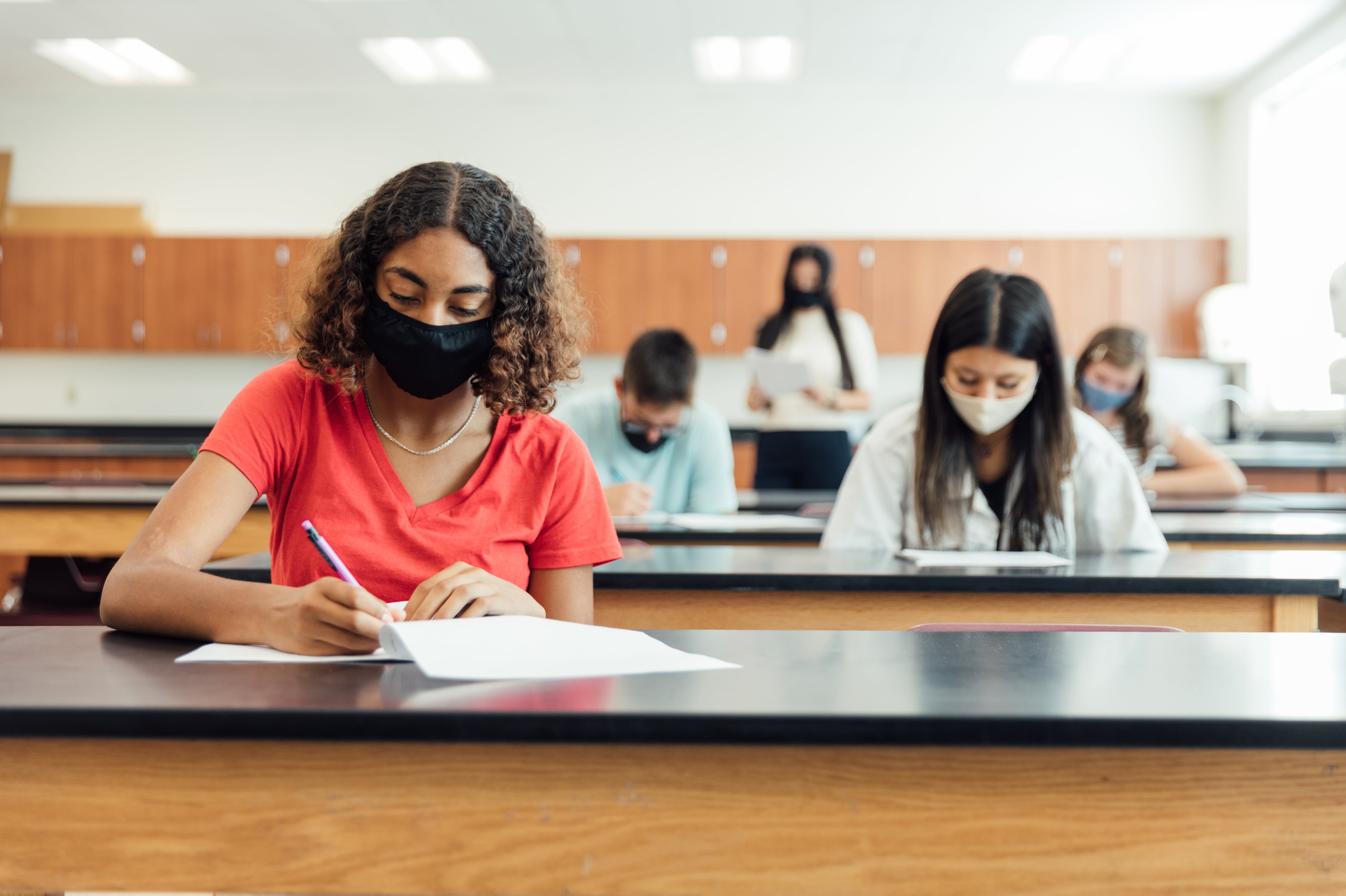 Masked high school students sit separated in rows of lab desks in a classroom.