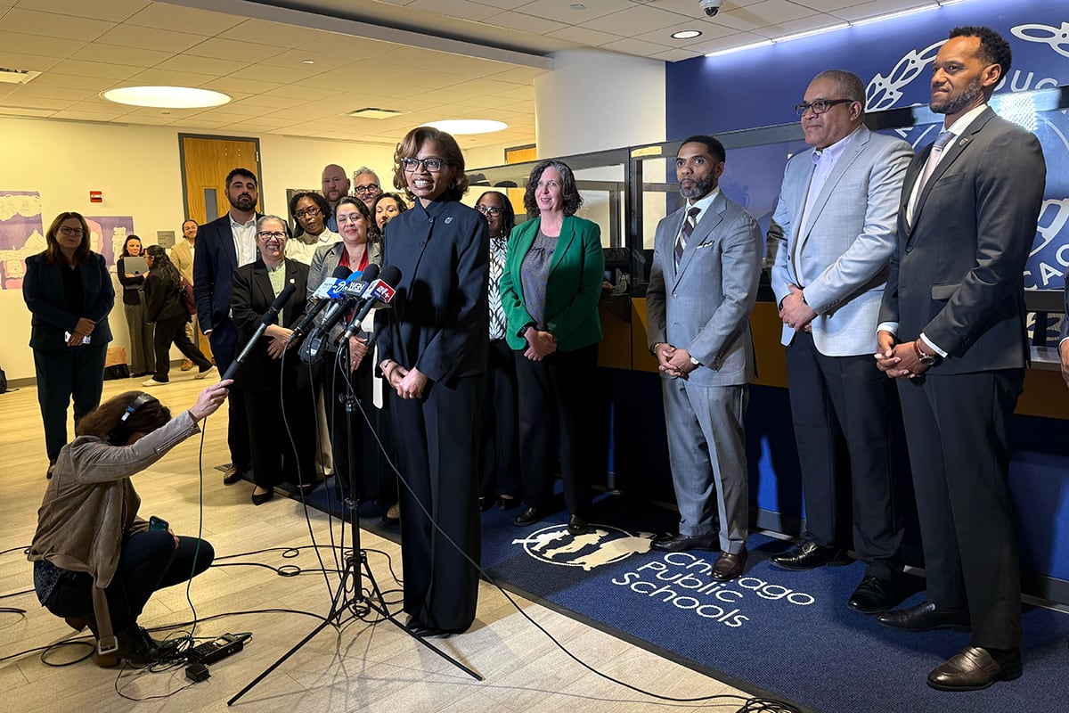 A woman in a dark suit stands at a microphone in front of a group of people.