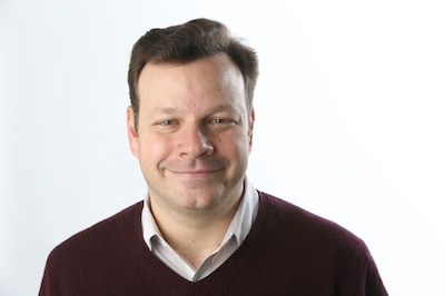 Headshot of a white man with brown hair wearing a white shirt and a black sweater.