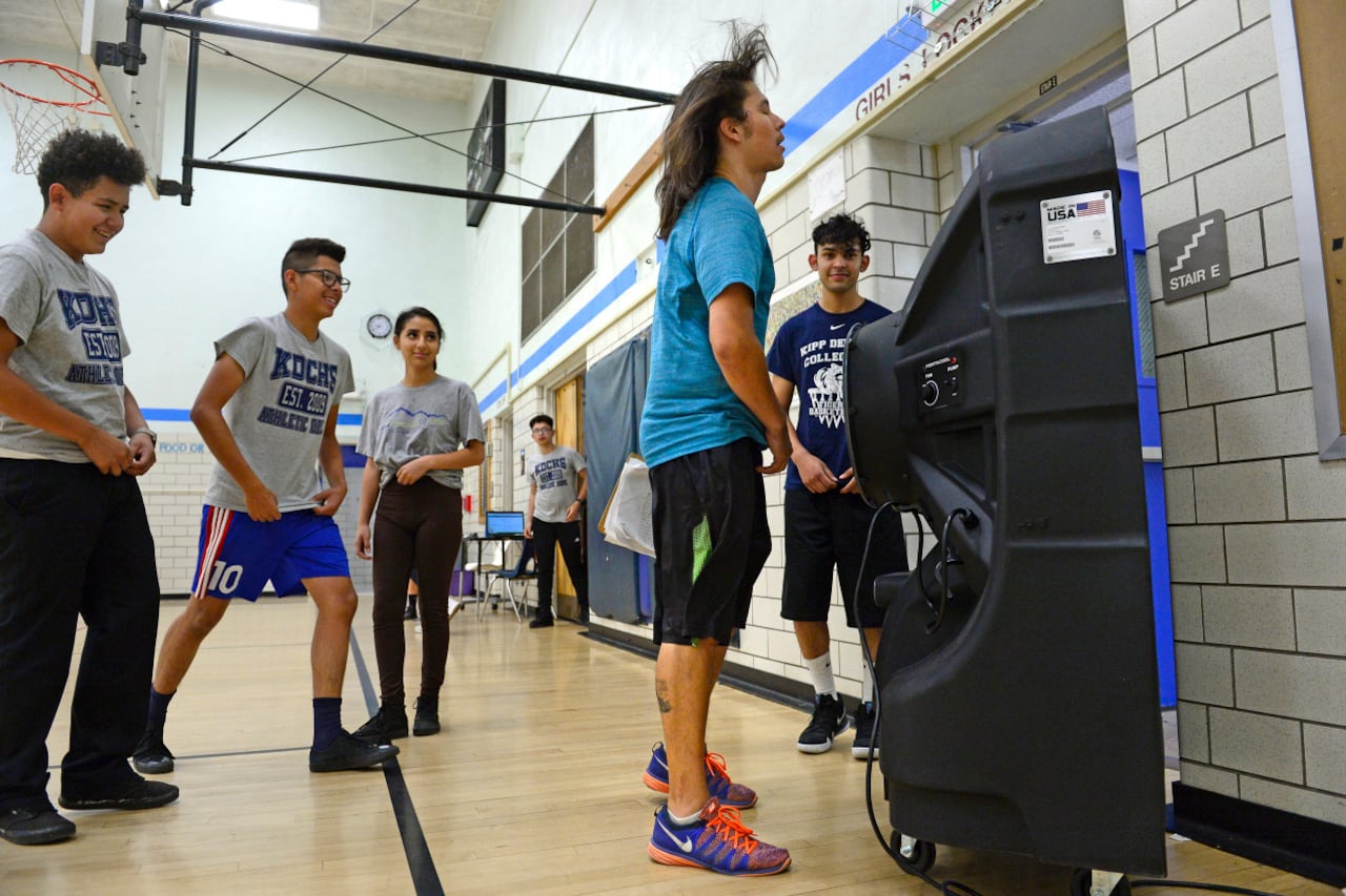 Students and a teacher in a gym stand around a portable air conditioning system. The air blows back the teacher’s hair.