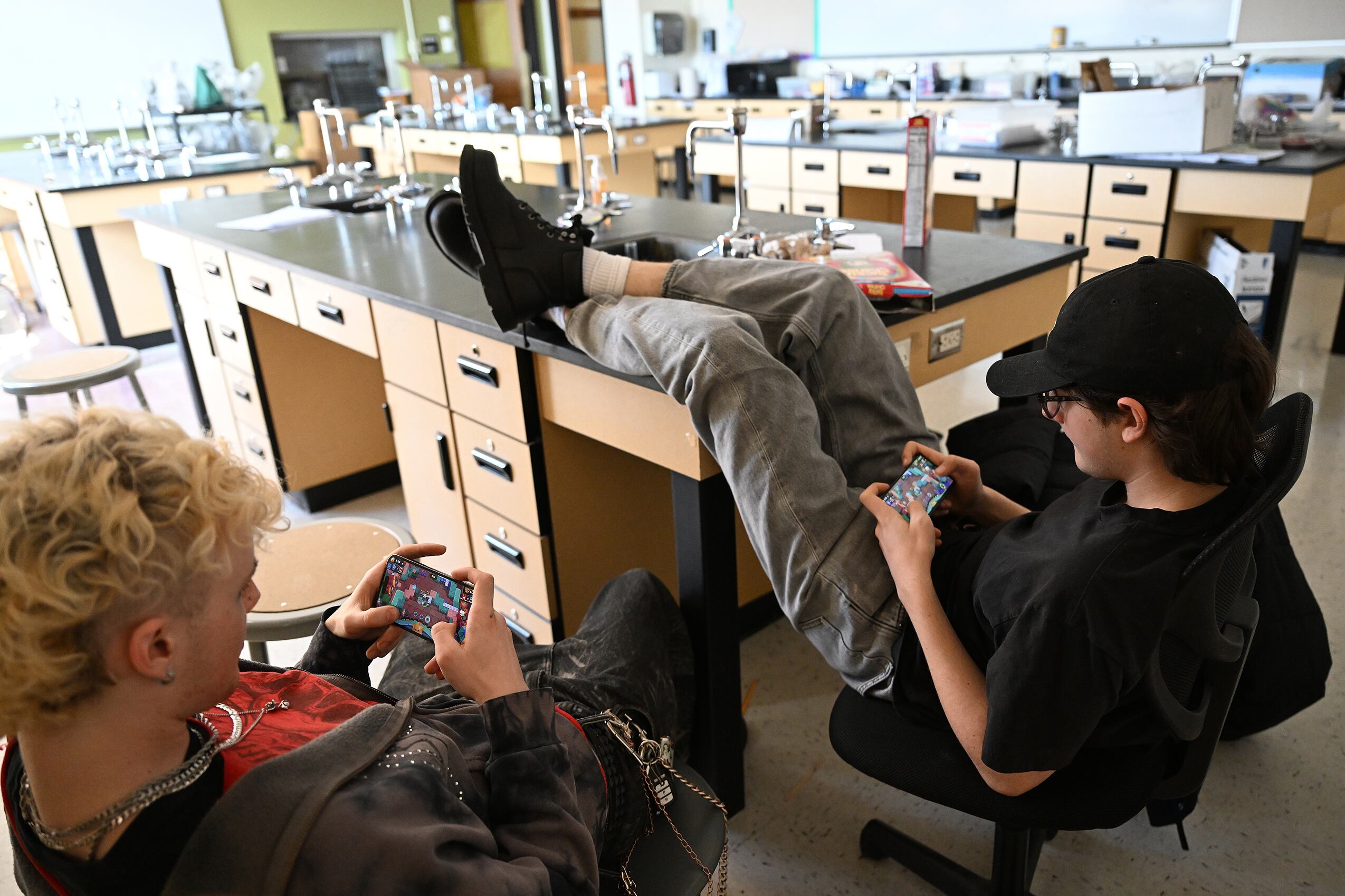 A photograph of two high school students sitting in a classroom on their cellphones.