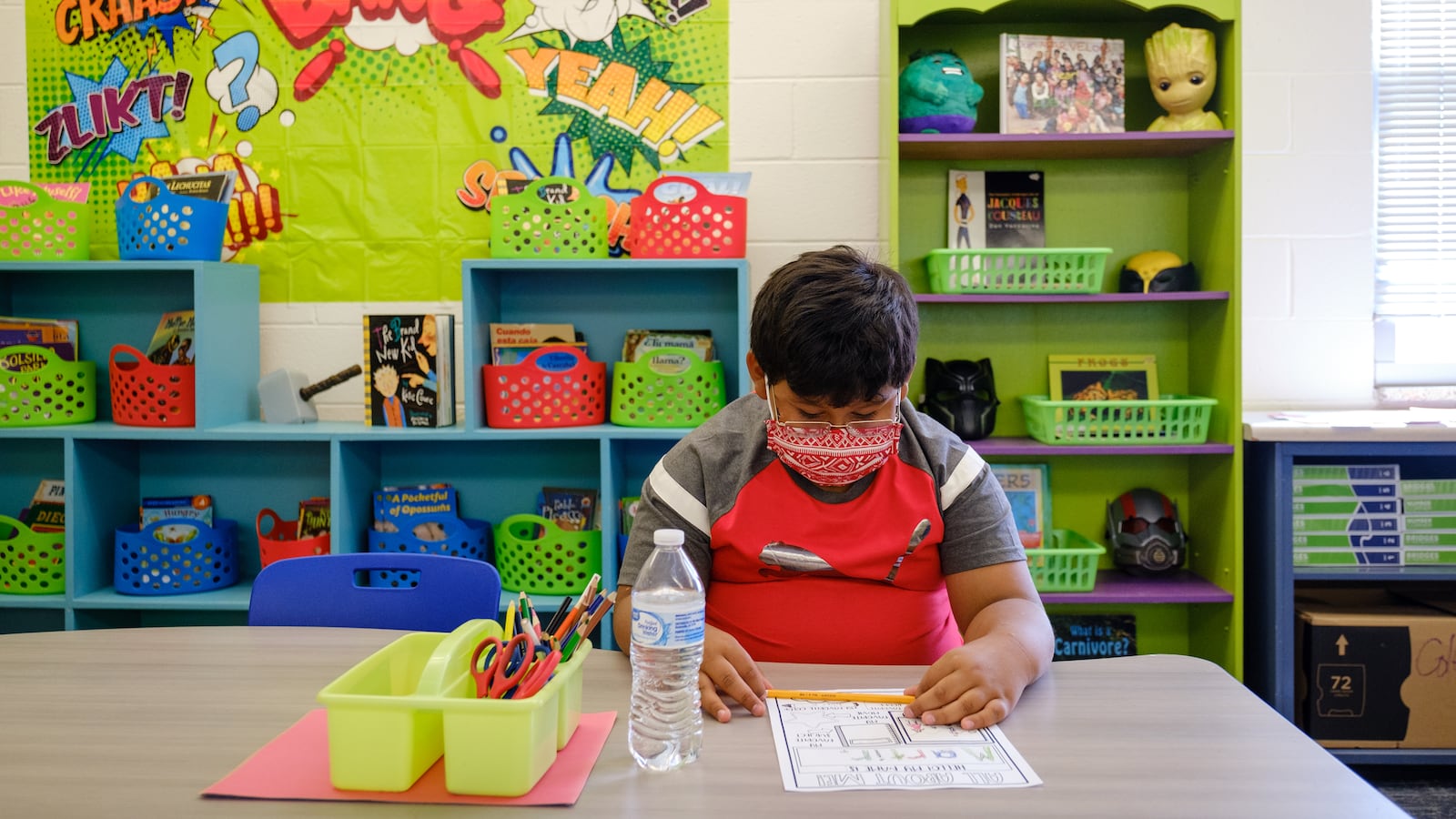 A student completes an introductory worksheet about himself for the first day of school. There are several colorful signs and bins on the wall behind him.