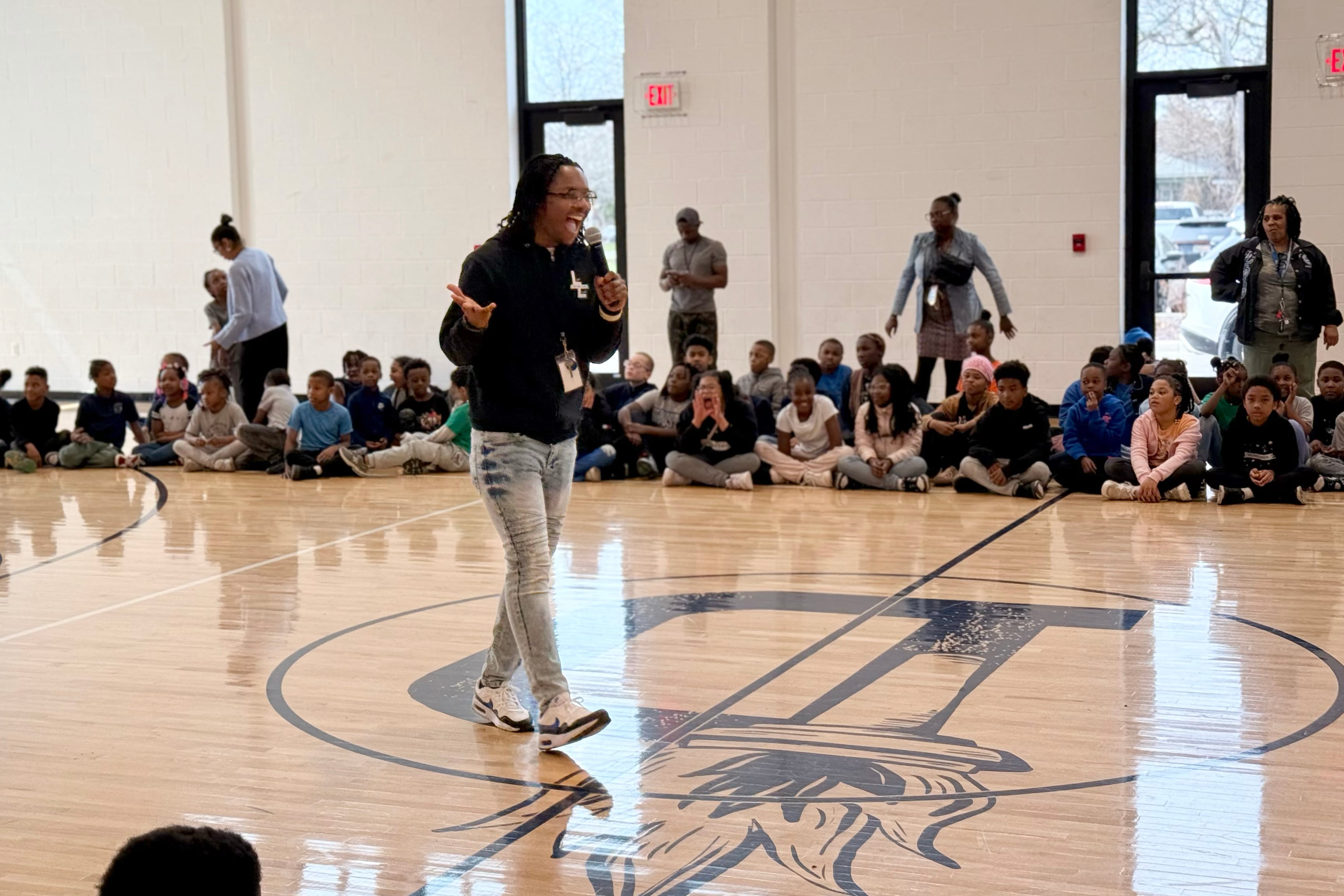 A Black man wearing a sweater and jeans speaks from the middle of a gymnasium floor holding a microphone with a row of students and educators in the background.