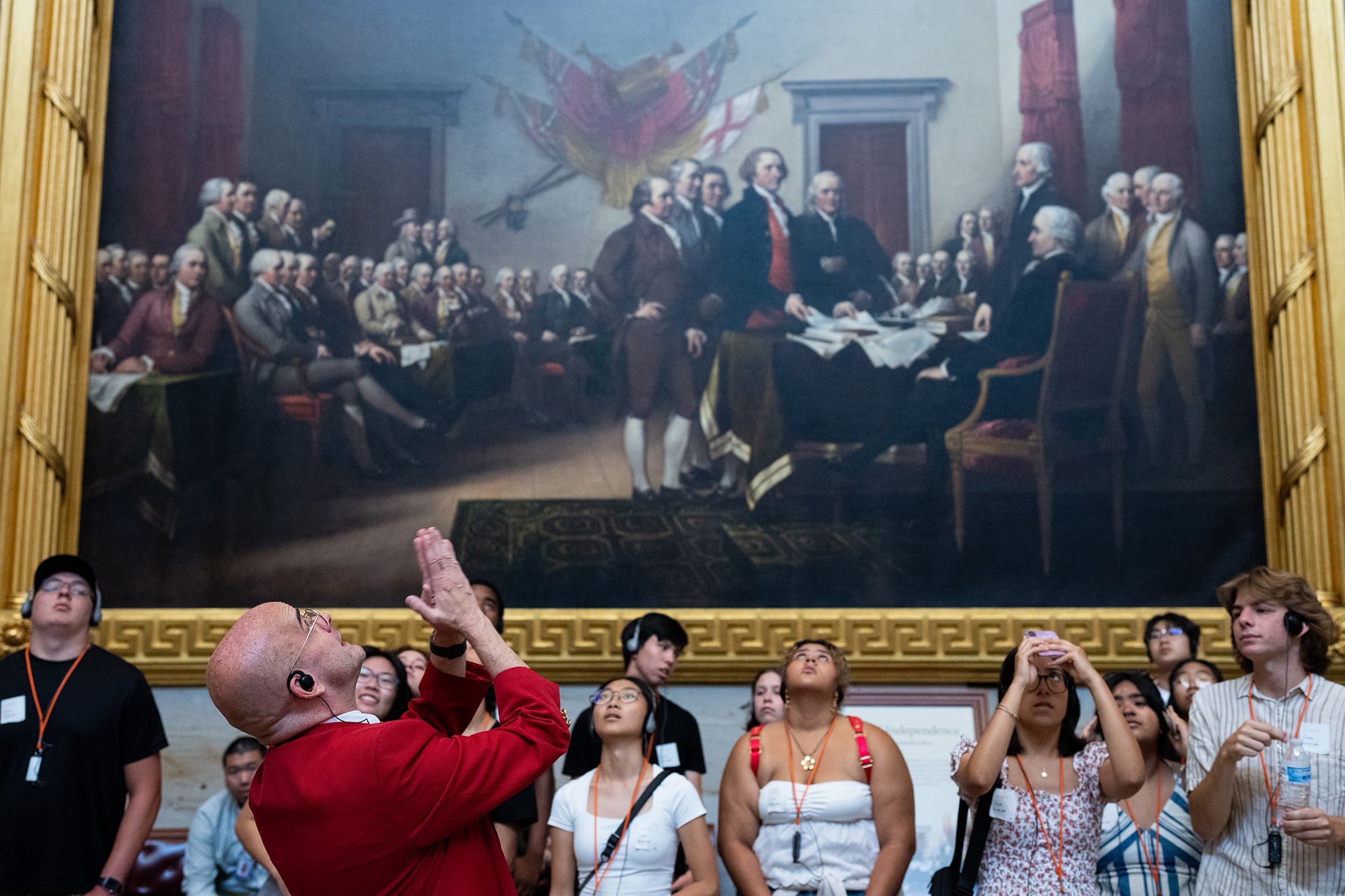A photograph of a large painting depicting the signing of the Declaration of Independence hung on a wall with a large group of tourists stare up at the ceiling.
