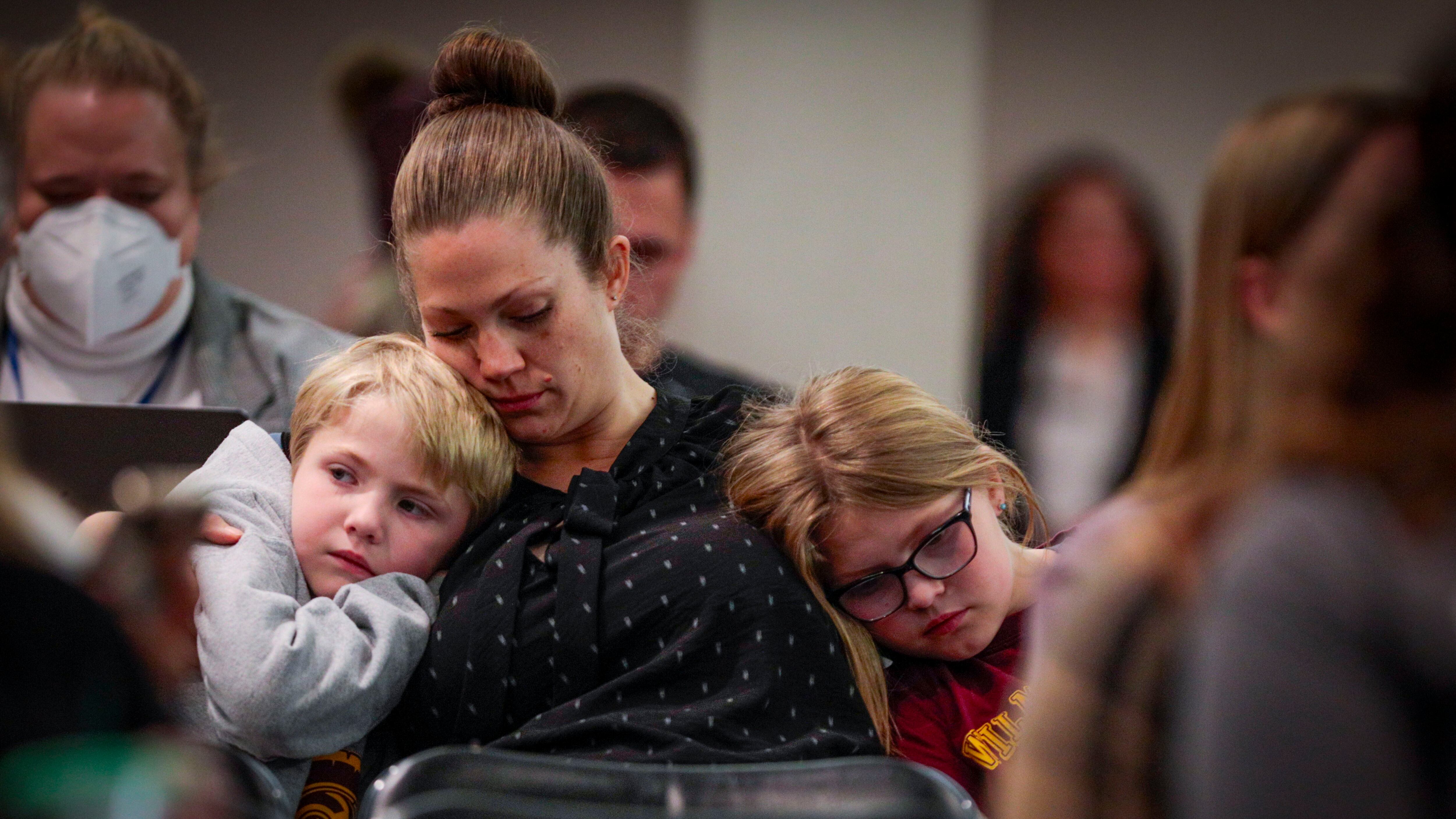 A woman with her hair pulled up in a bun comforts two children who nestle against her. She’s sitting in a crowded room surrounded by people.