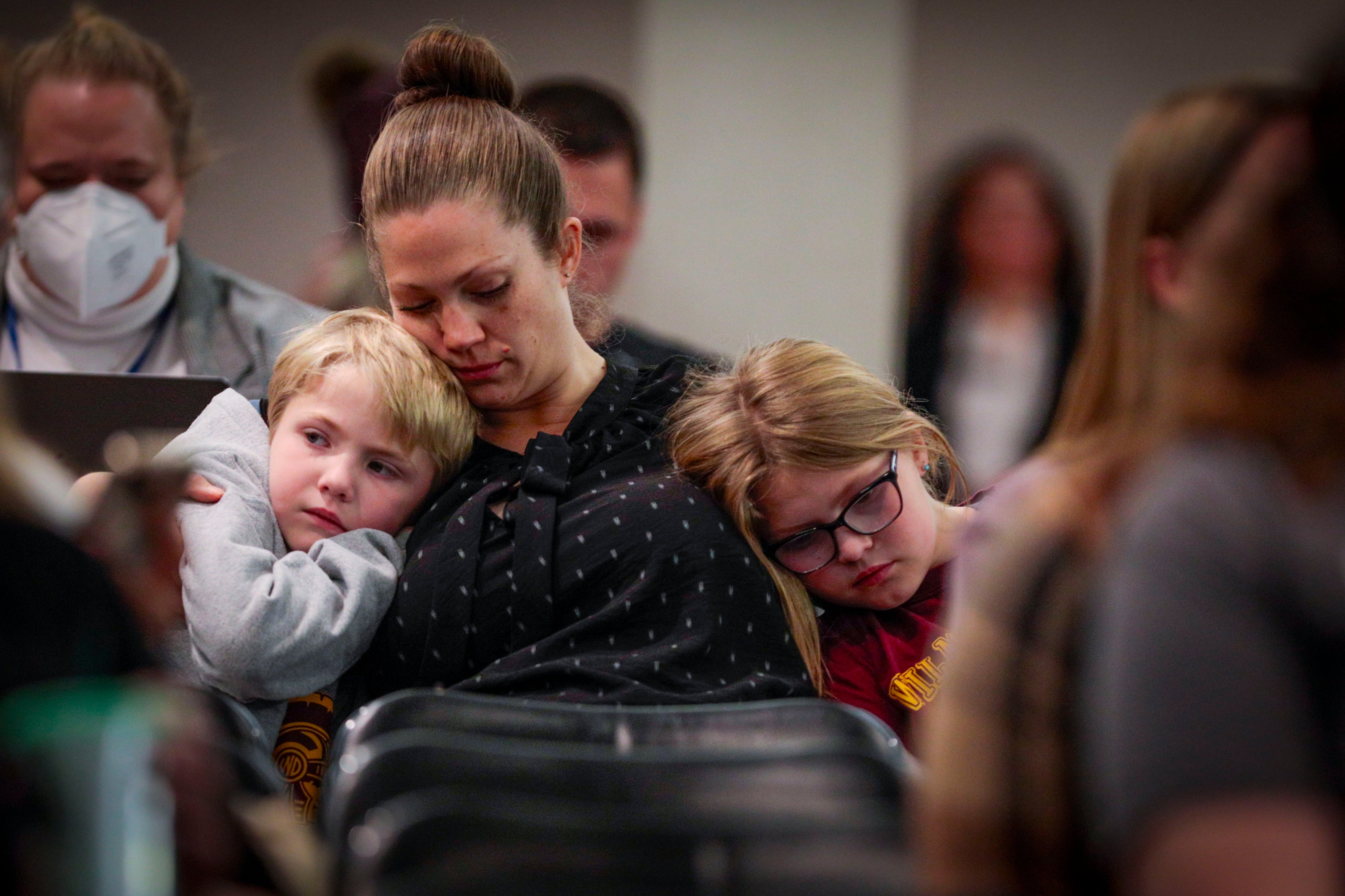 A woman with her hair pulled up in a bun comforts two children who nestle against her. She’s sitting in a crowded room surrounded by people.