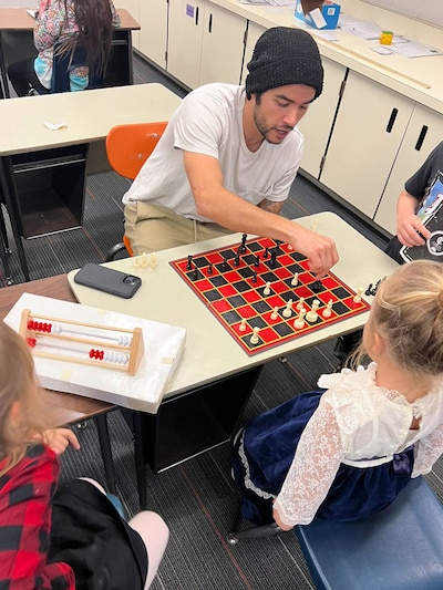 An adult man plays chess in a classroom against a young student while two other students stand nearby.