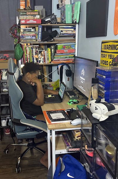 A photograph of a young Black student working on a laptop in their room.