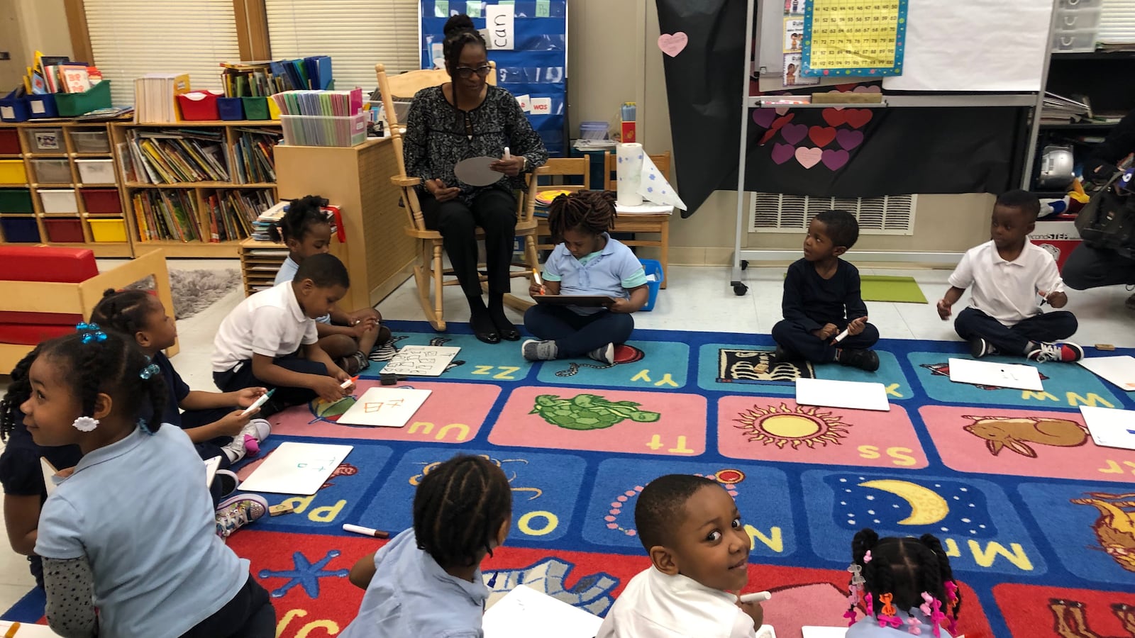A classroom full of young students sitting on a colorful rug in a circle while a teacher sits in a chair reading from a book.