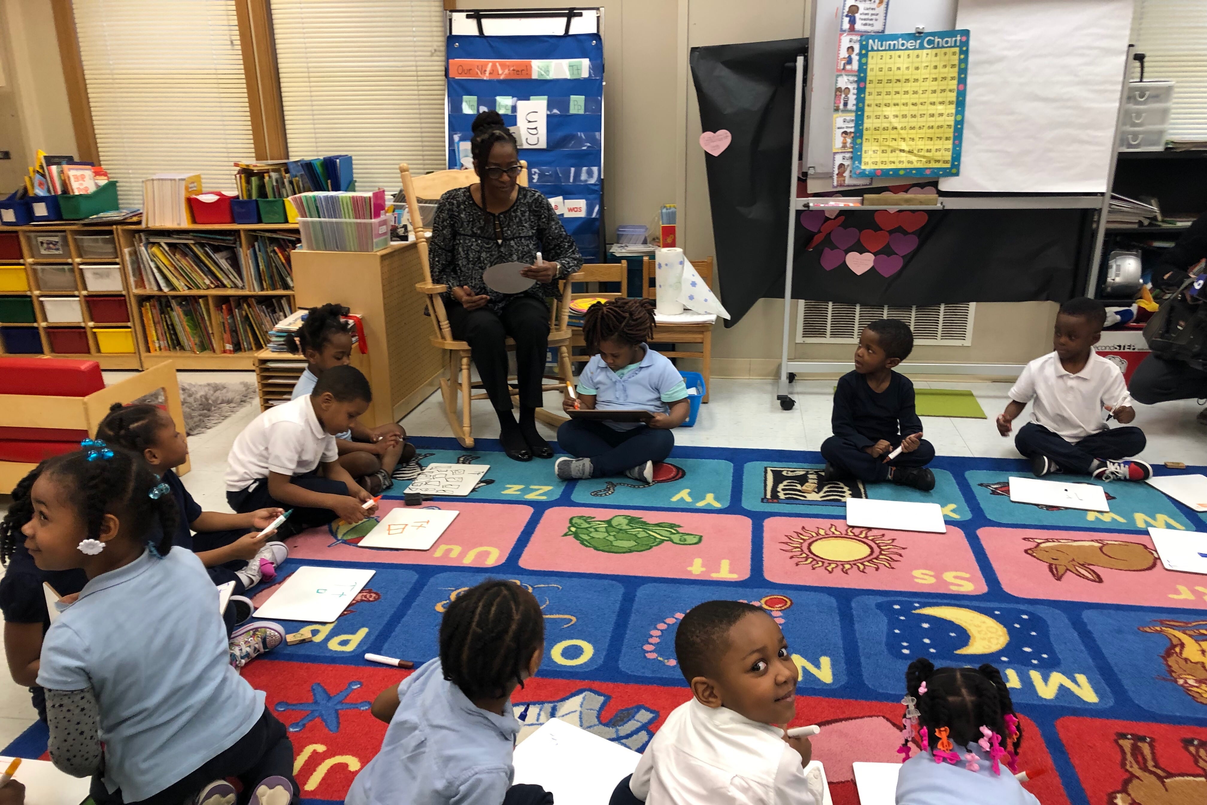 A classroom full of young students sitting on a colorful rug in a circle while a teacher sits in a chair reading from a book.