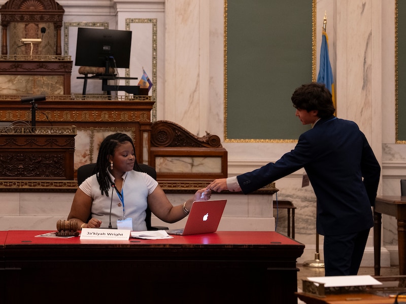 A photograph of a Black teenage woman receiving a note from another teenage man at the desk.