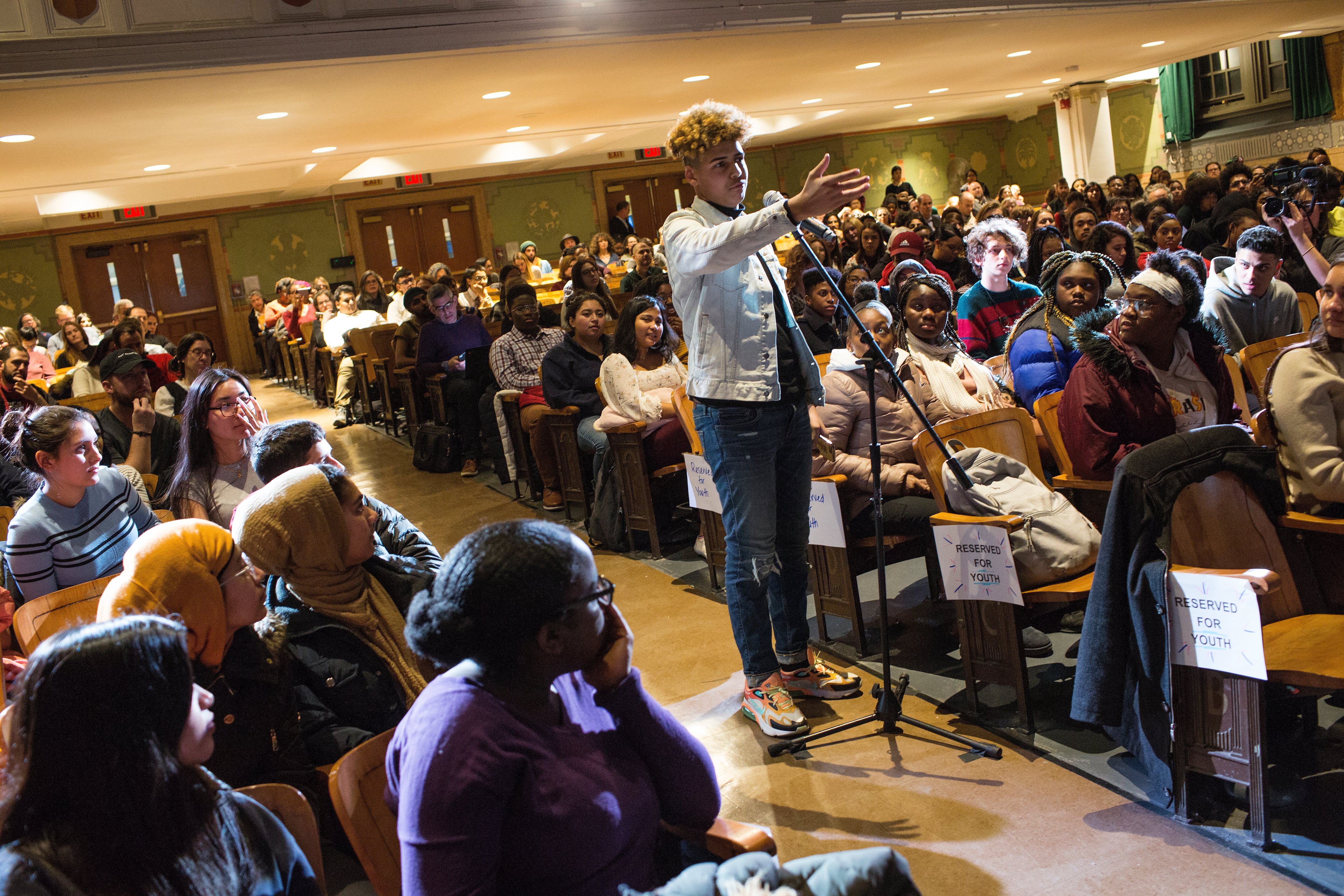A teen in a white denim jacket stands at a microphone in a school auditorium as the crowd in their seats look on.