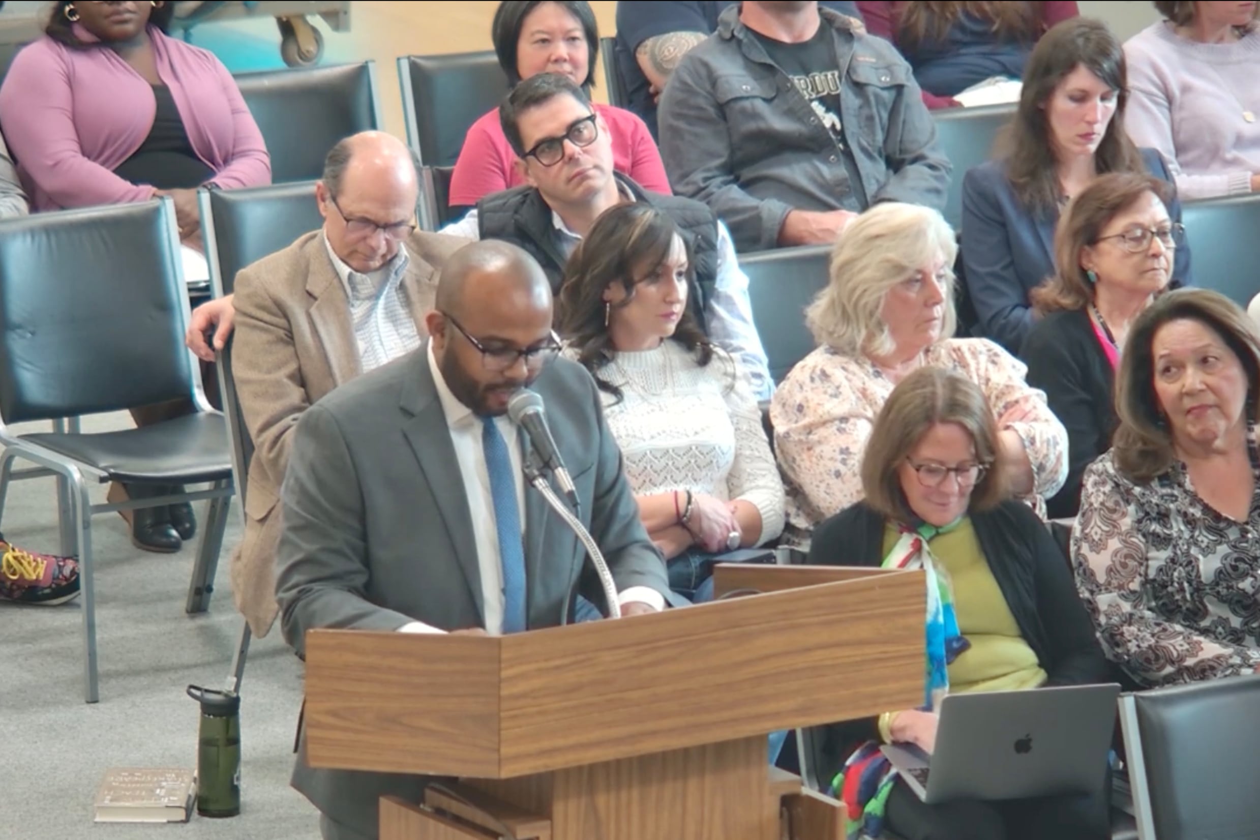 A Black man in a suit speaks from a microphone with a group of people sitting in rows of seats in the background.