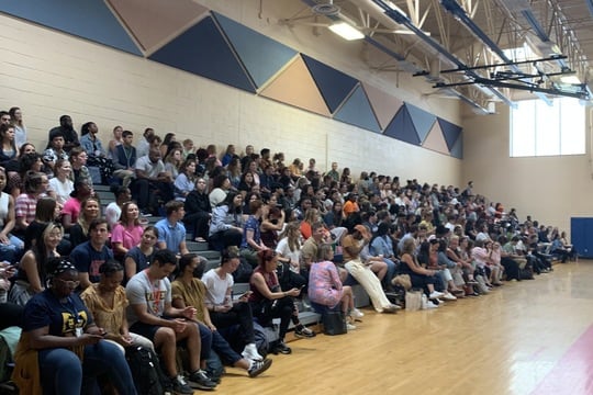 People sit on bleachers in a school gymnasium.