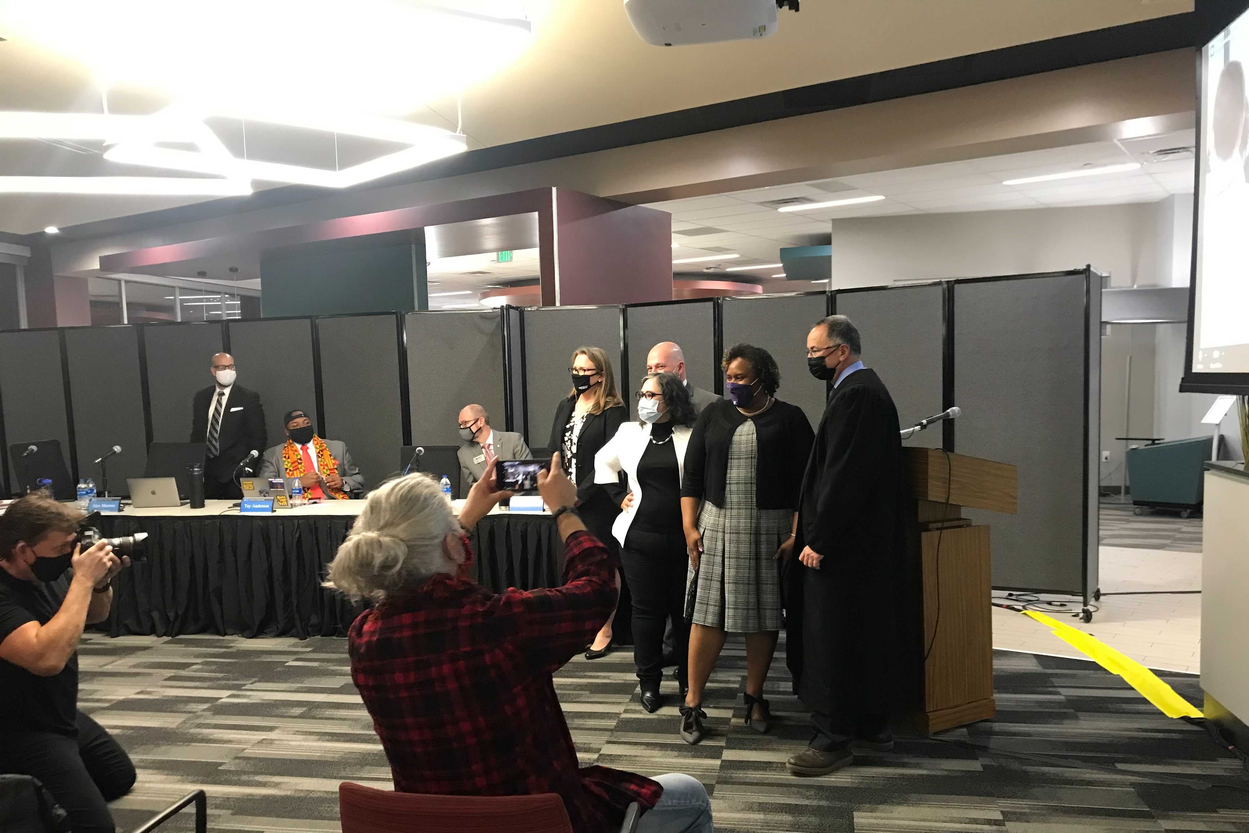 Denver school board members pose for a photo in front of a podium. People in the audience hold up cell phones to take photos.