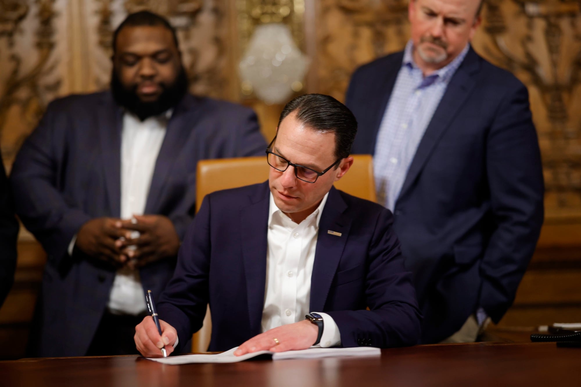 A man wearing a suit sits at a wooden table signing a piece of paper while two men in suits stand behind.