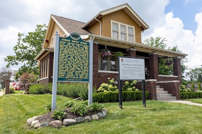 A photograph of a large brick house with a green historical sign in the lawn.
