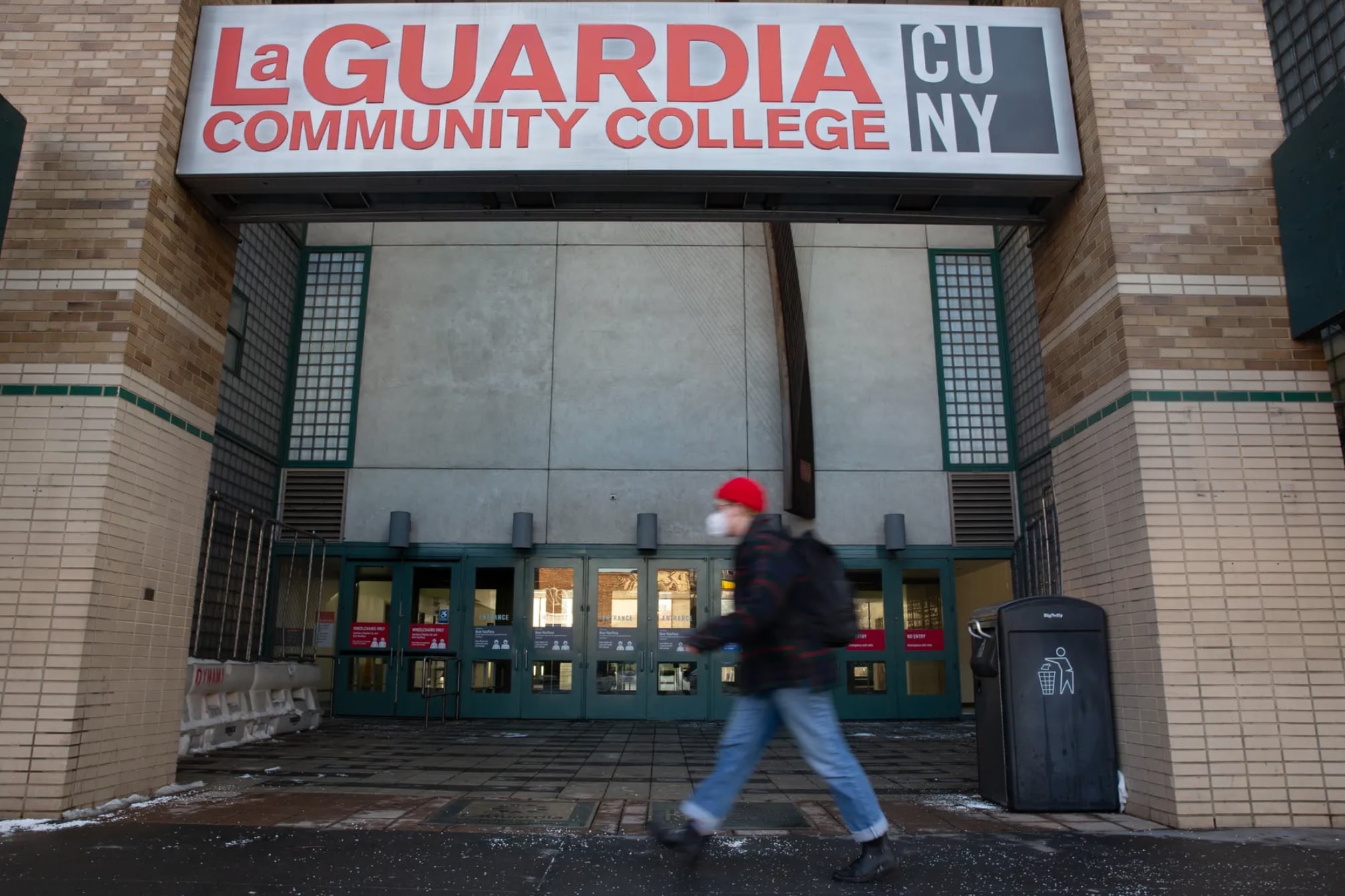 A person walks by the entrance of LaGuardia Community College, wearing a red beanie.