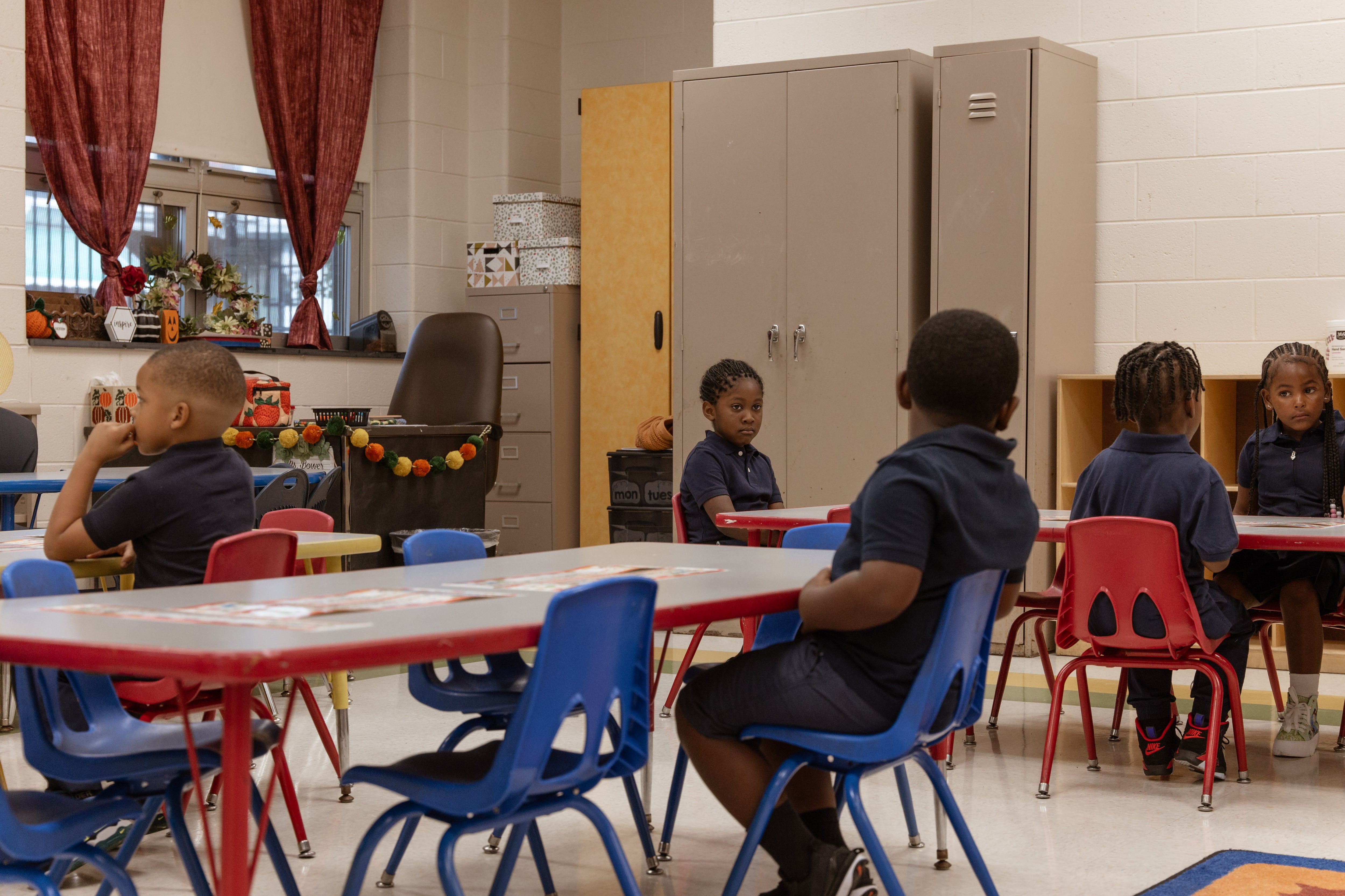 Young students in uniform sit at different tables inside a classroom.