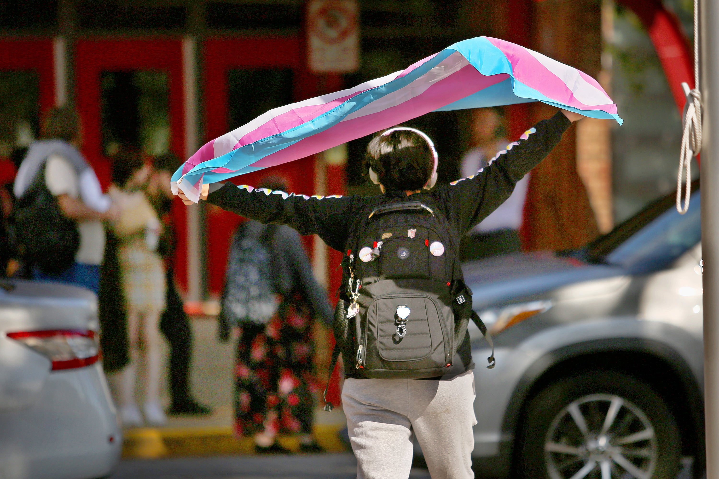 A high school student wearing a black backpack and holding a transgender flag above their head walks toward a building with people and cars in the background.