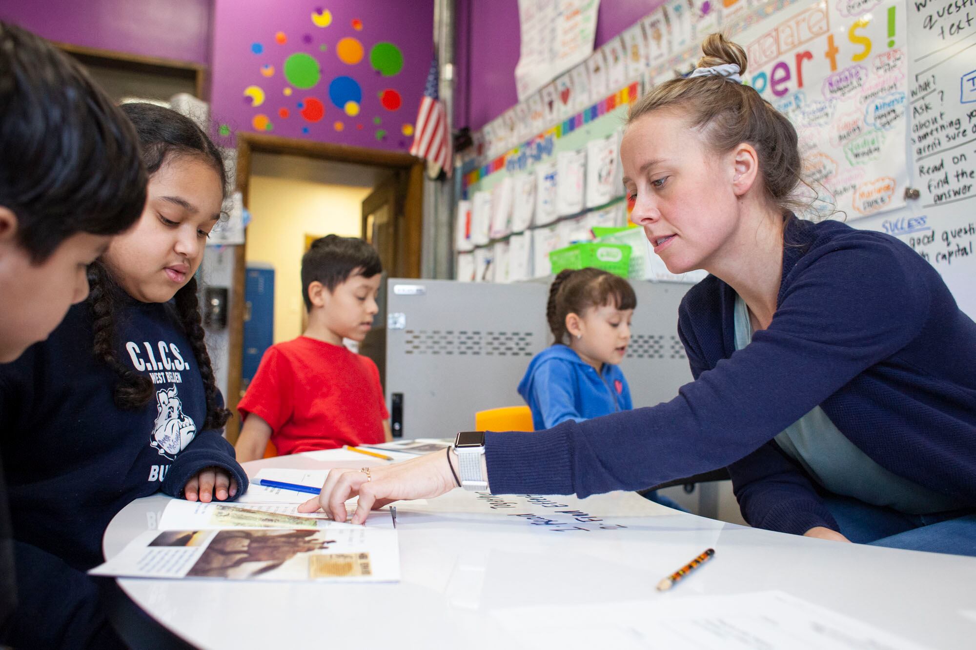 A photograph of a white teacher sitting at a desk with four young students in a colorful classroom.