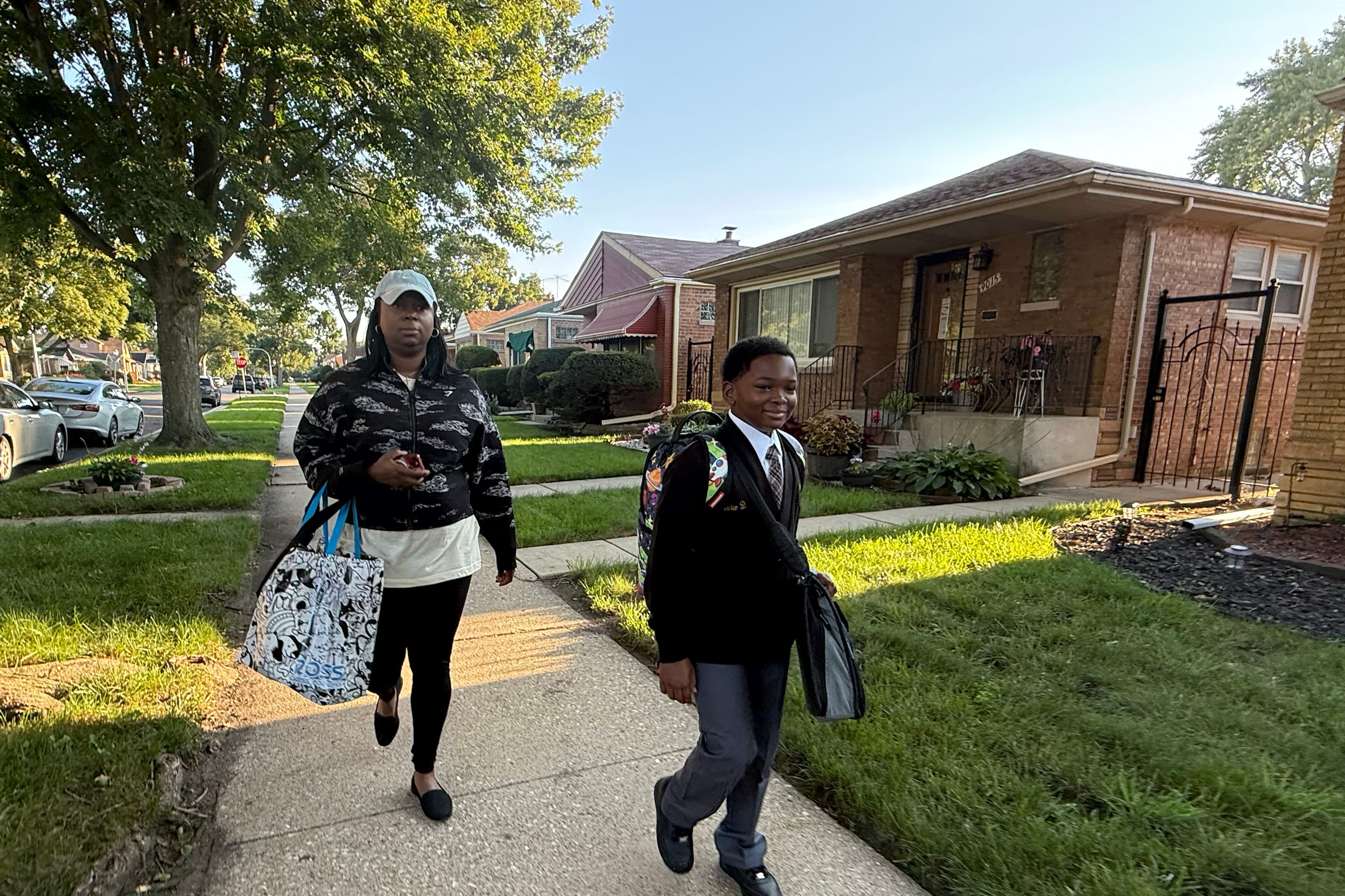 A Black mom and her son walk down a sidewalk on a sunny morning passing houses.