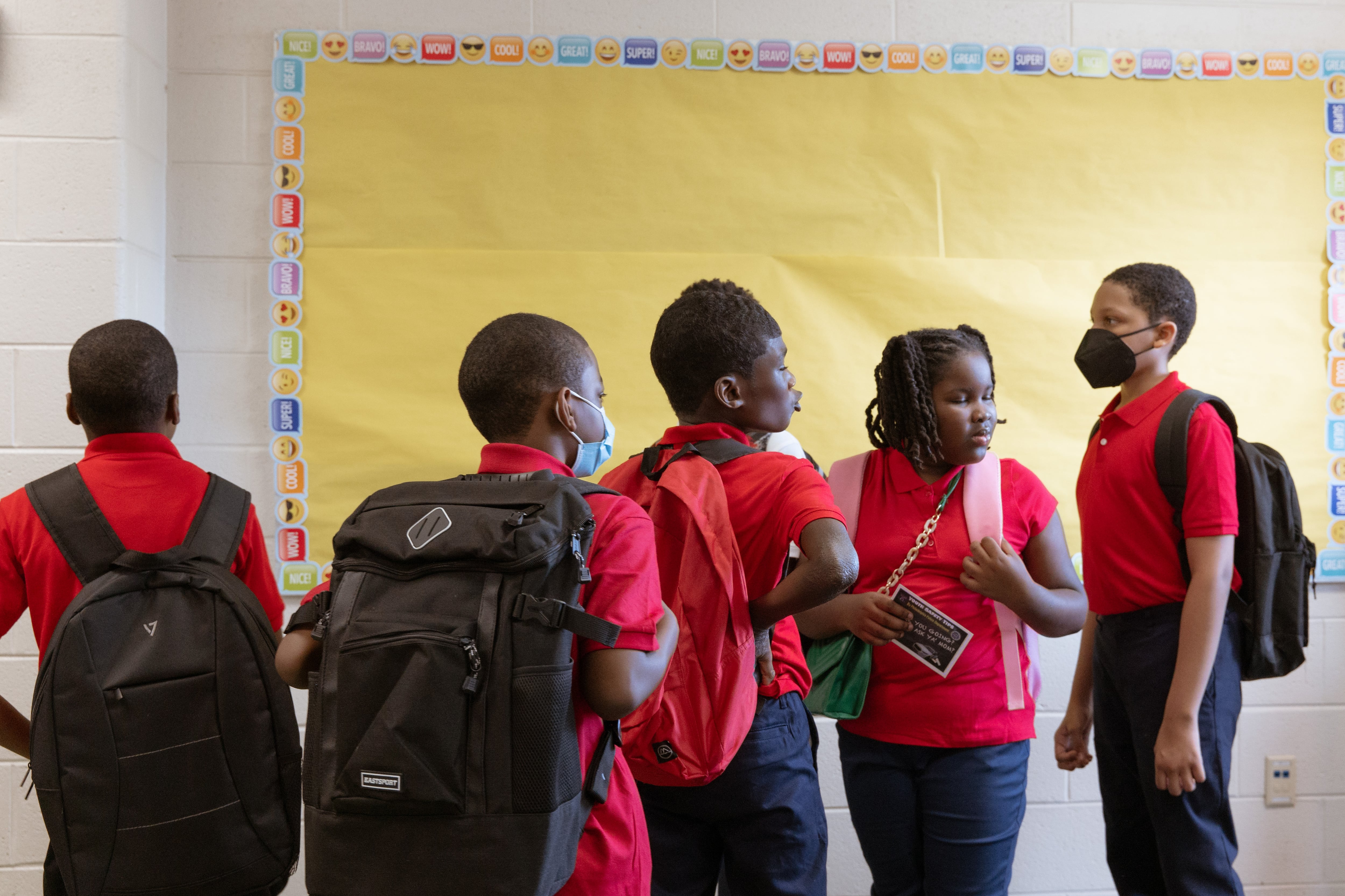 Five students wearing red and black uniforms stand in a hallway with their backpacks on and a giant yellow piece of paper on the wall in the background.