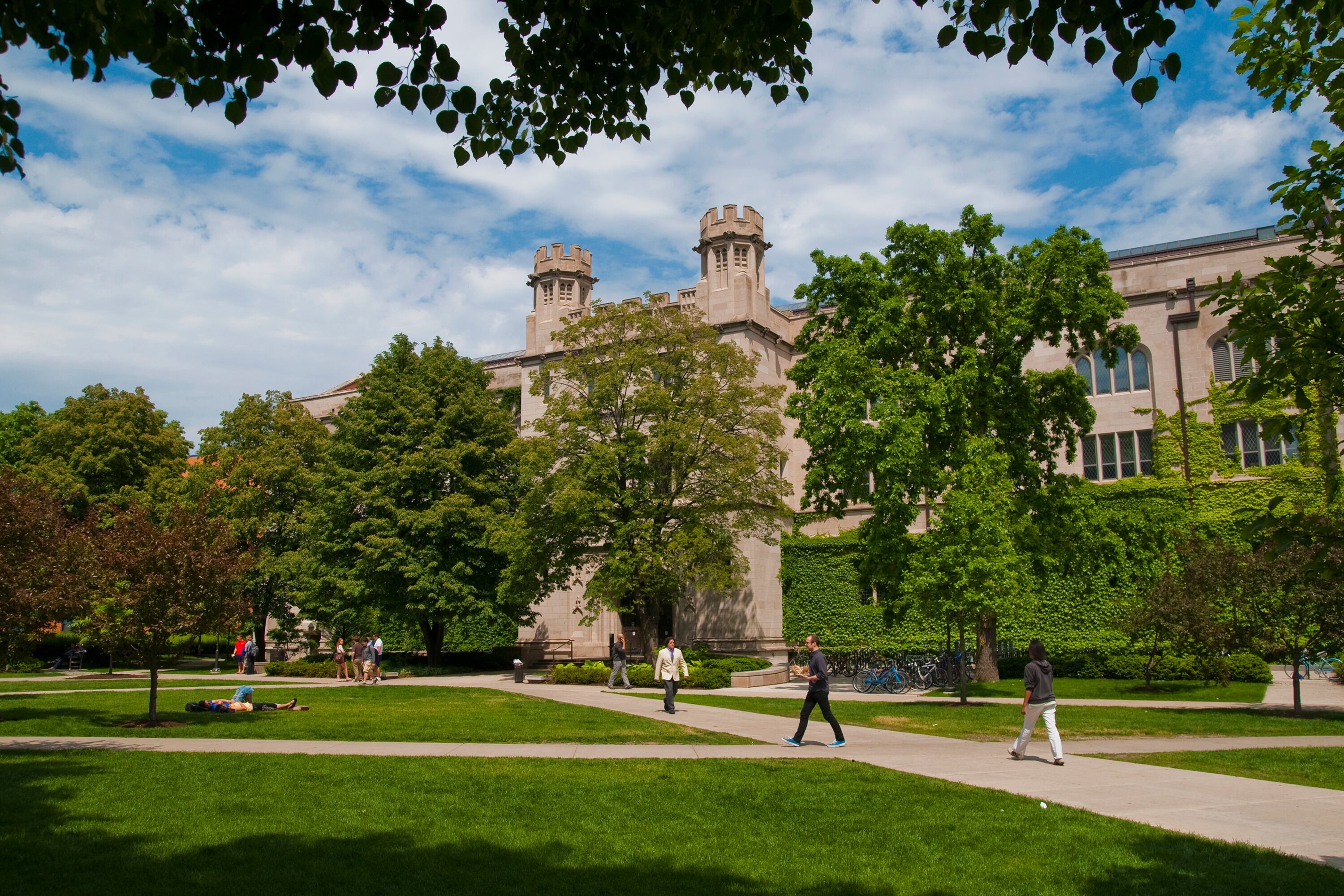 People walk on paths in front of trees and a building.