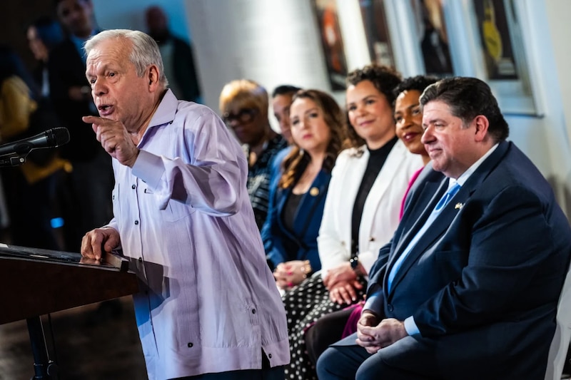 A man in a light blue shirt speaks and points while at a podium. A row of others sit behind him.