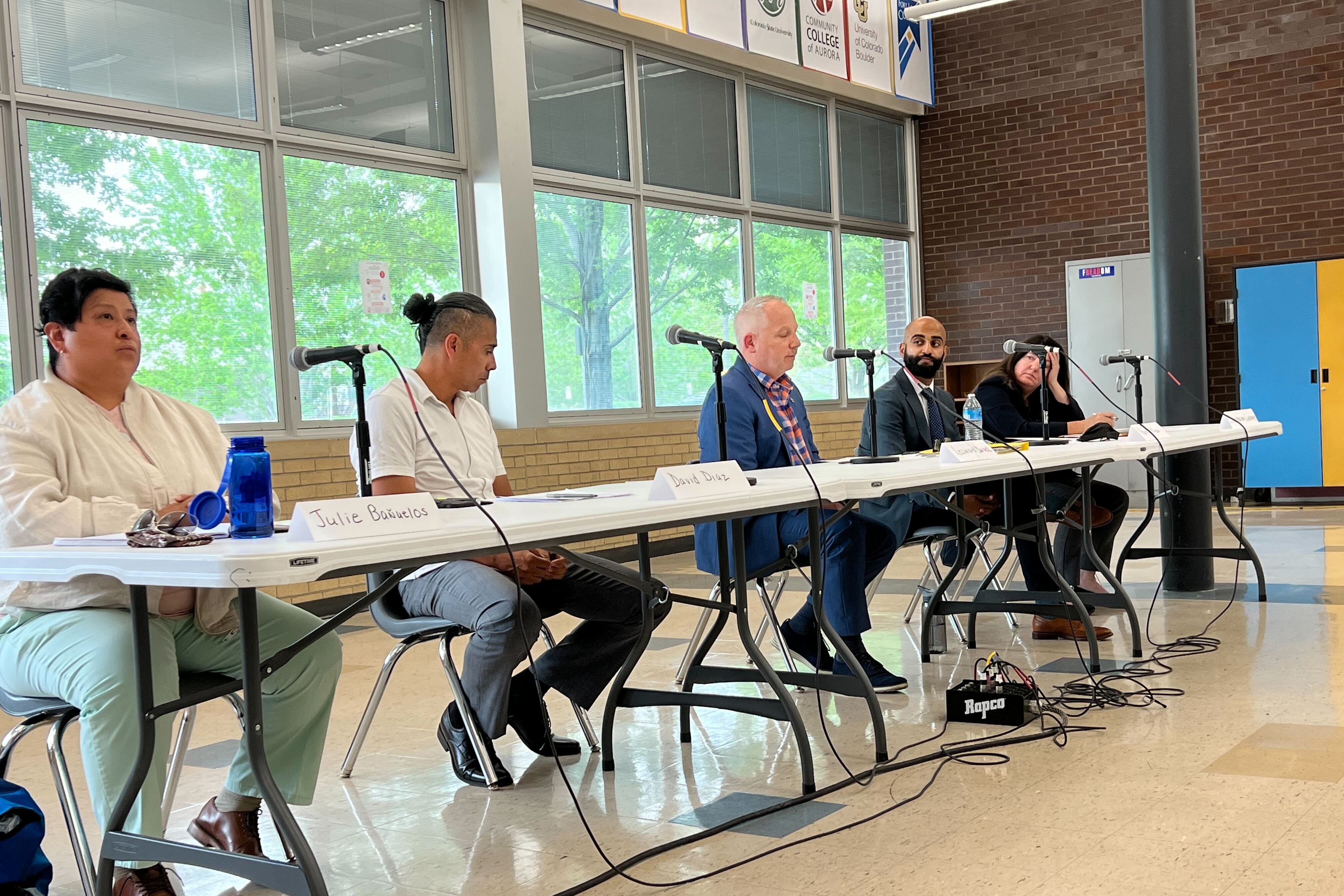 Five candidates sit at a long folding table in a high school cafeteria. Each candidate has a microphone in front of them.