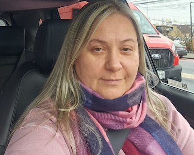 A woman with long blond hair and wearing a pink shirt and colorful scarf looks directly at the camera from inside of a car with a red car in the background.