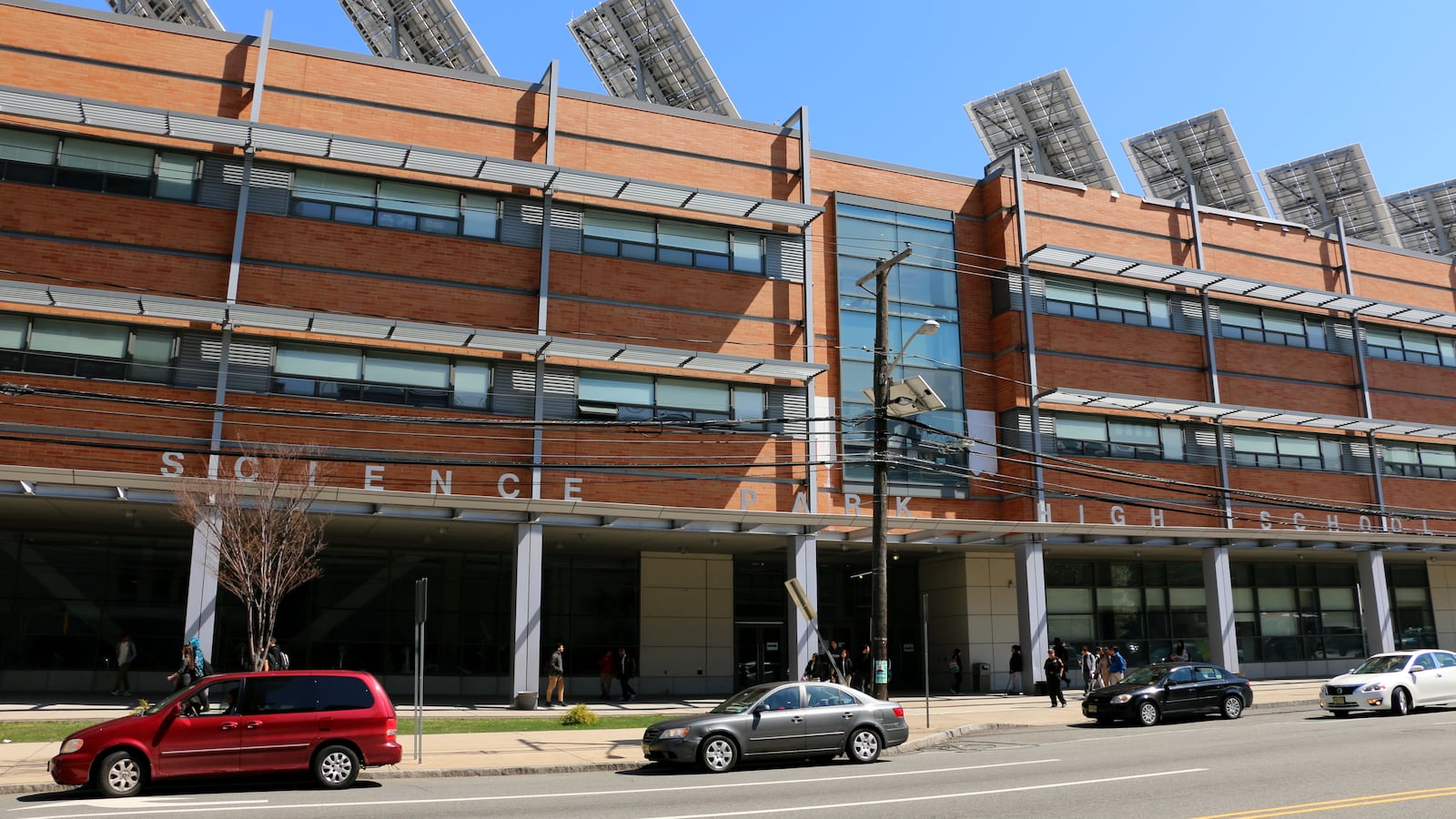 A red building with the words Science Park High School is seen along with three cars parked outside on the street.