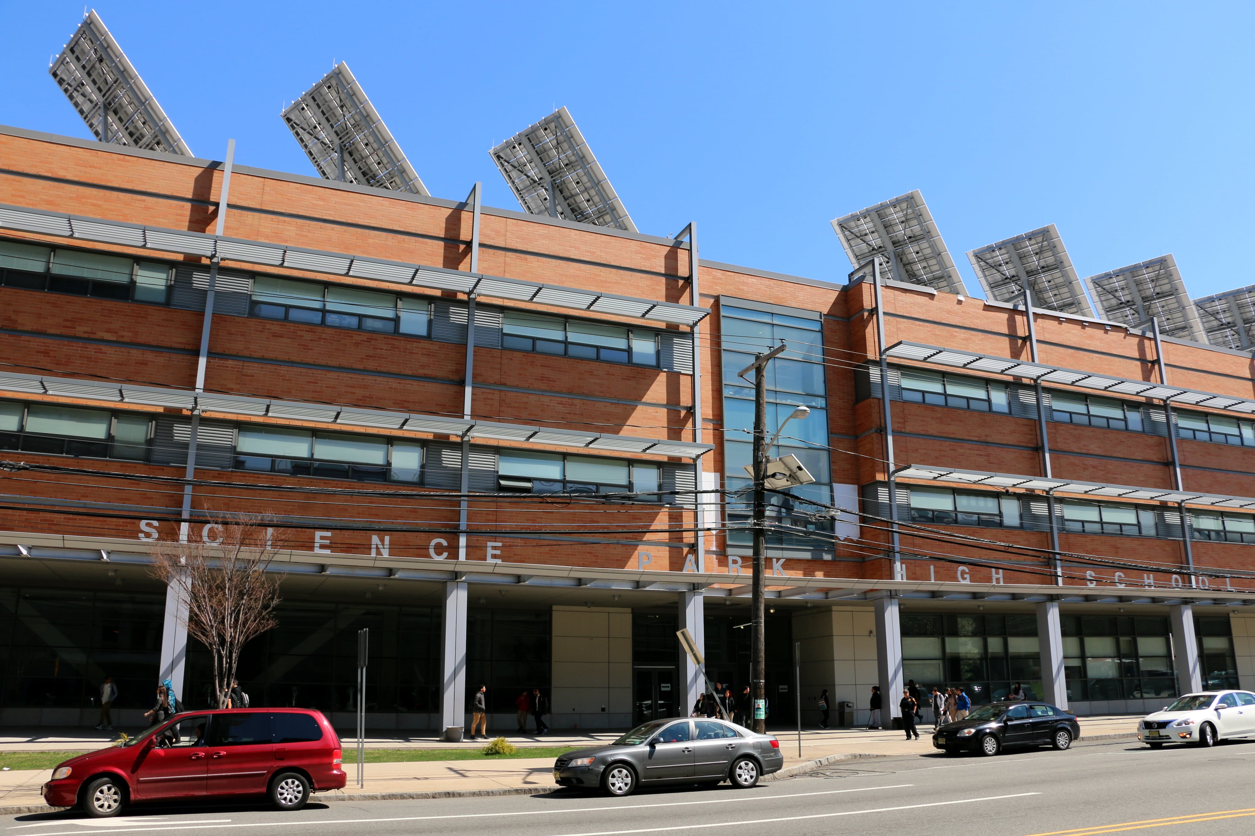 A red building with the words Science Park High School is seen along with three cars parked outside on the street.