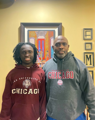 A boy wearing a red sweater stands next to his father who is wearing a grey sweater both smile and look at the camera. A painting in the background hangs on a yellow wall.