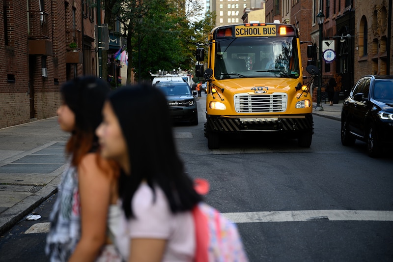A photograph of a yellow school bus in the background while two high school students walk across the street in the foreground.