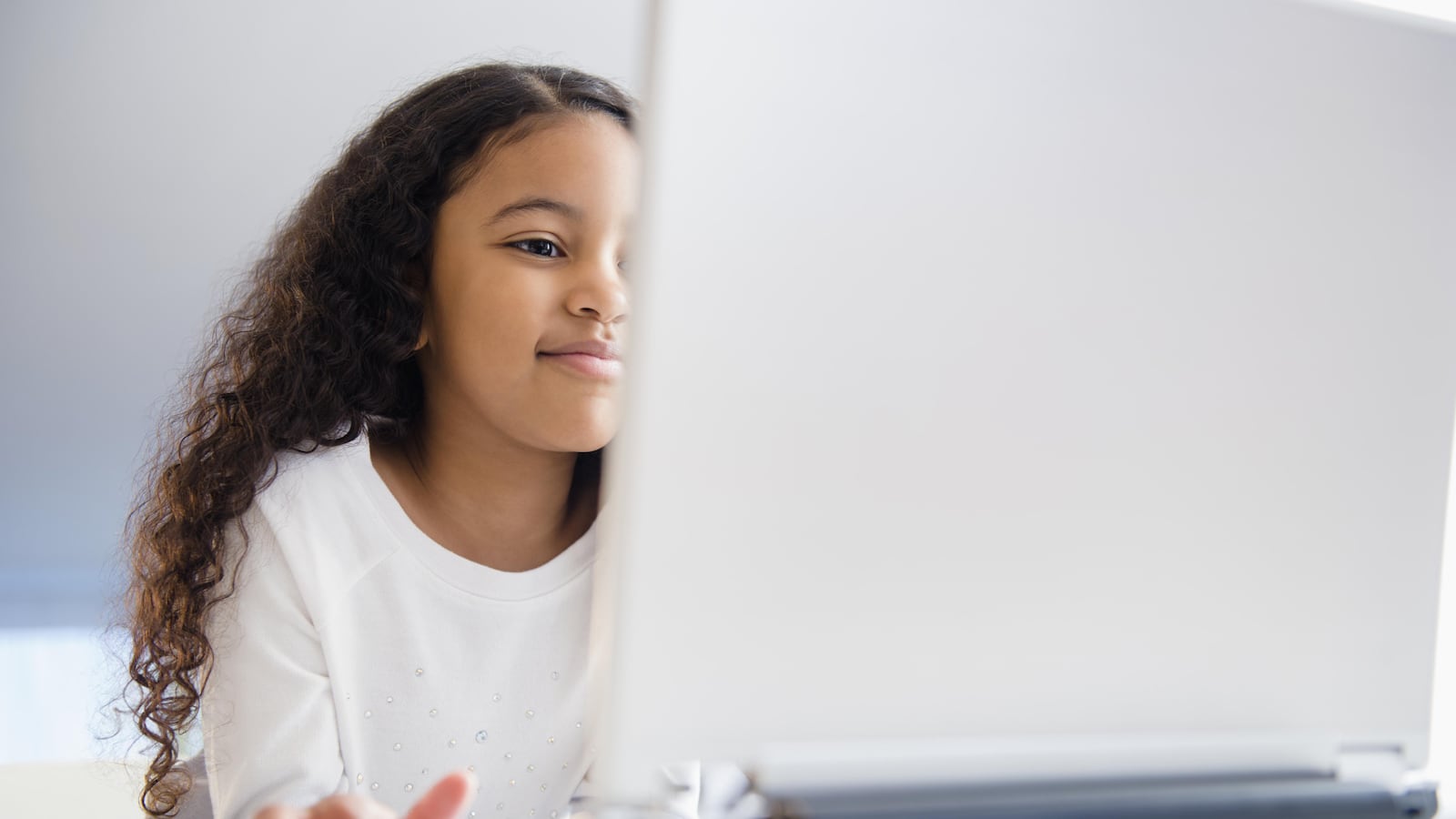 A young girl with long hair wears a white shirt. She works on a laptop computer.