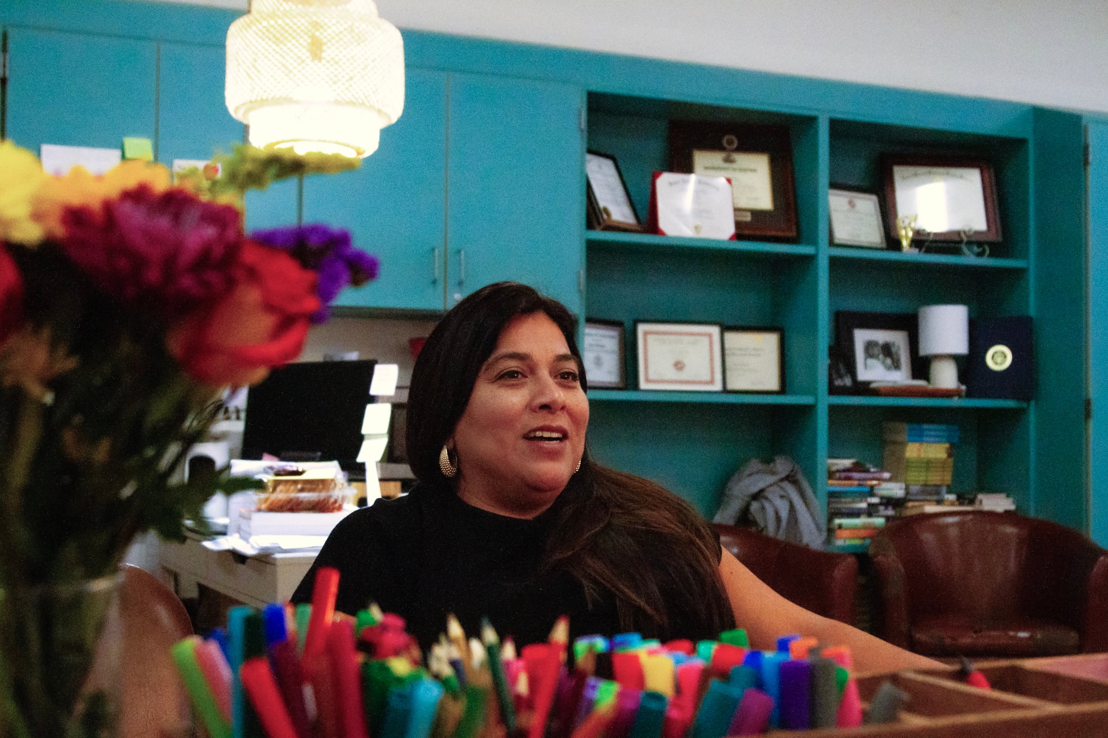 A woman in a black table sits at a desk in front of bookshelves.