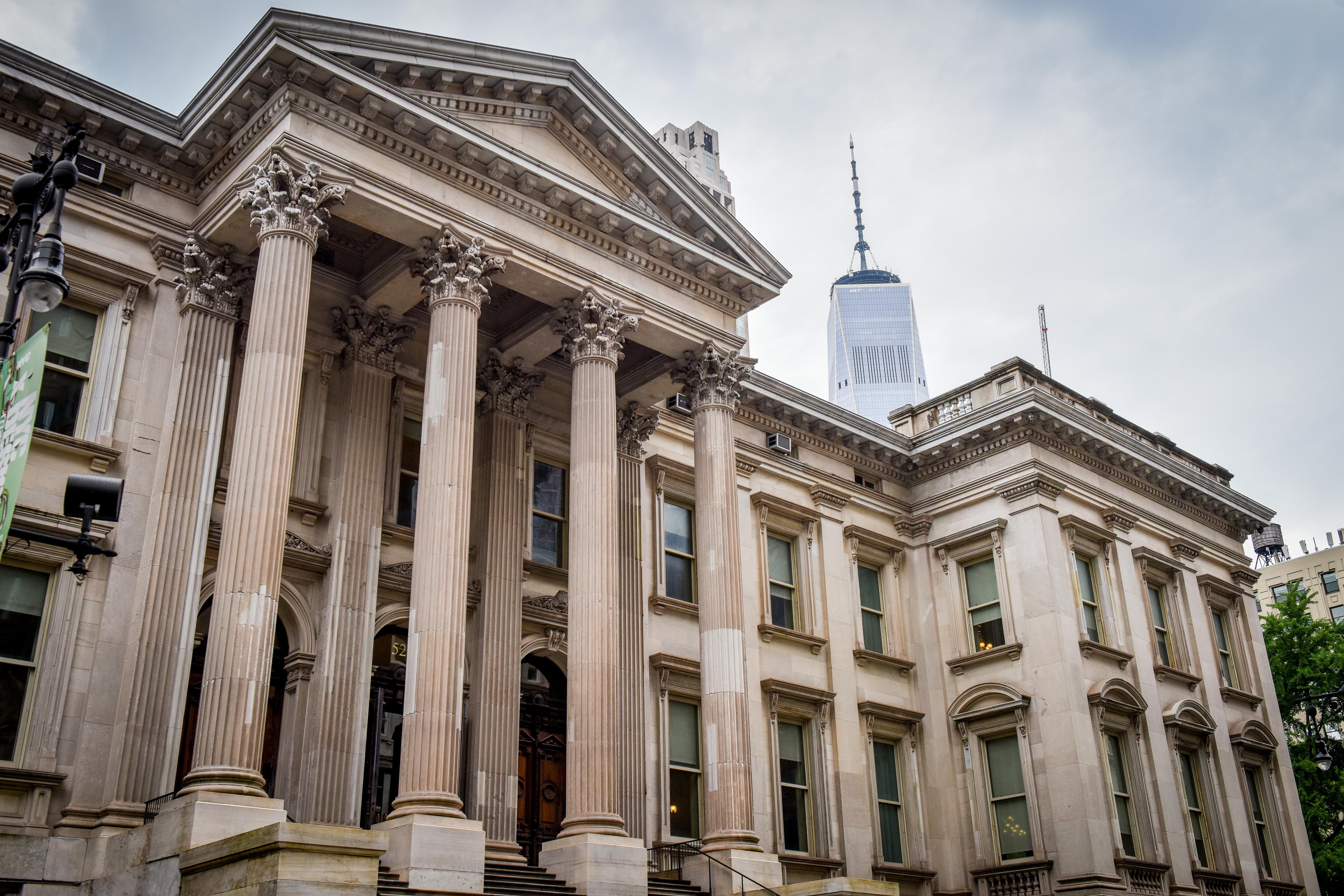 A white marble building with large columns with the One World Trade Center and a cloudy sky in the background.
